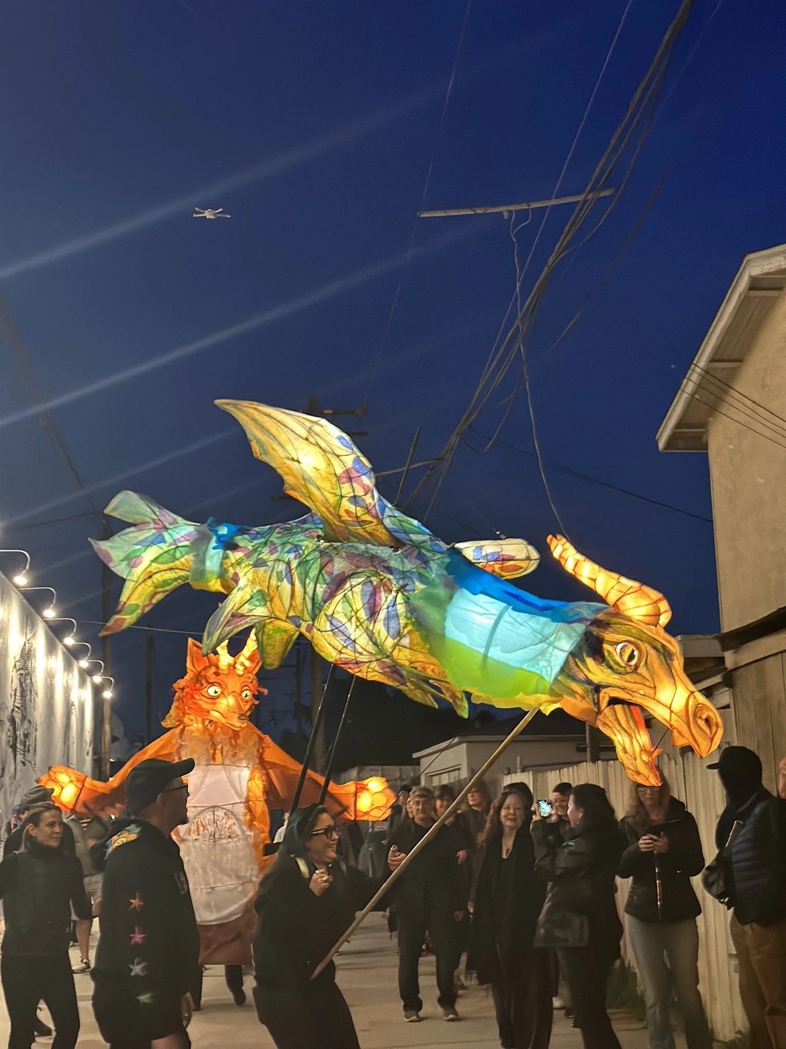People participating in a night festival parade, carrying colorful dragon and lion lantern sculptures, with onlookers watching along a street under a dark evening sky.