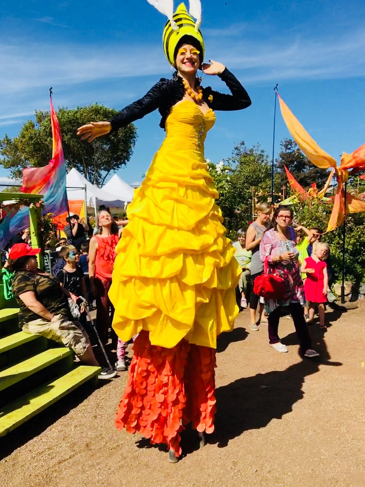 Performer on stilts dressed as a bee, wearing a yellow dress and large bee hat, at outdoor festival with colorful flags and crowd.