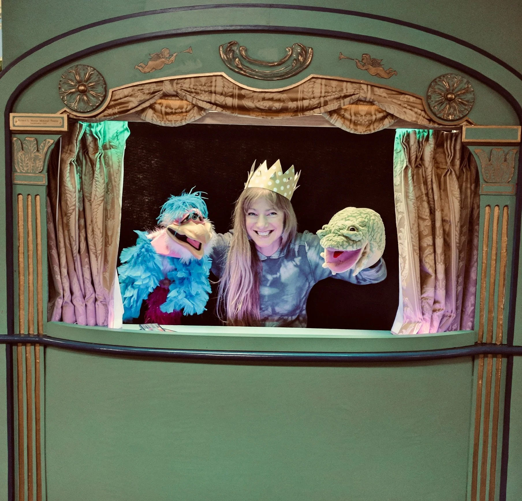 Woman with long hair wearing a crown, smiling, flanked by two puppet characters with dinosaur and bird appearances, at a theater stage with green and gold ornate decorations and pink curtains.