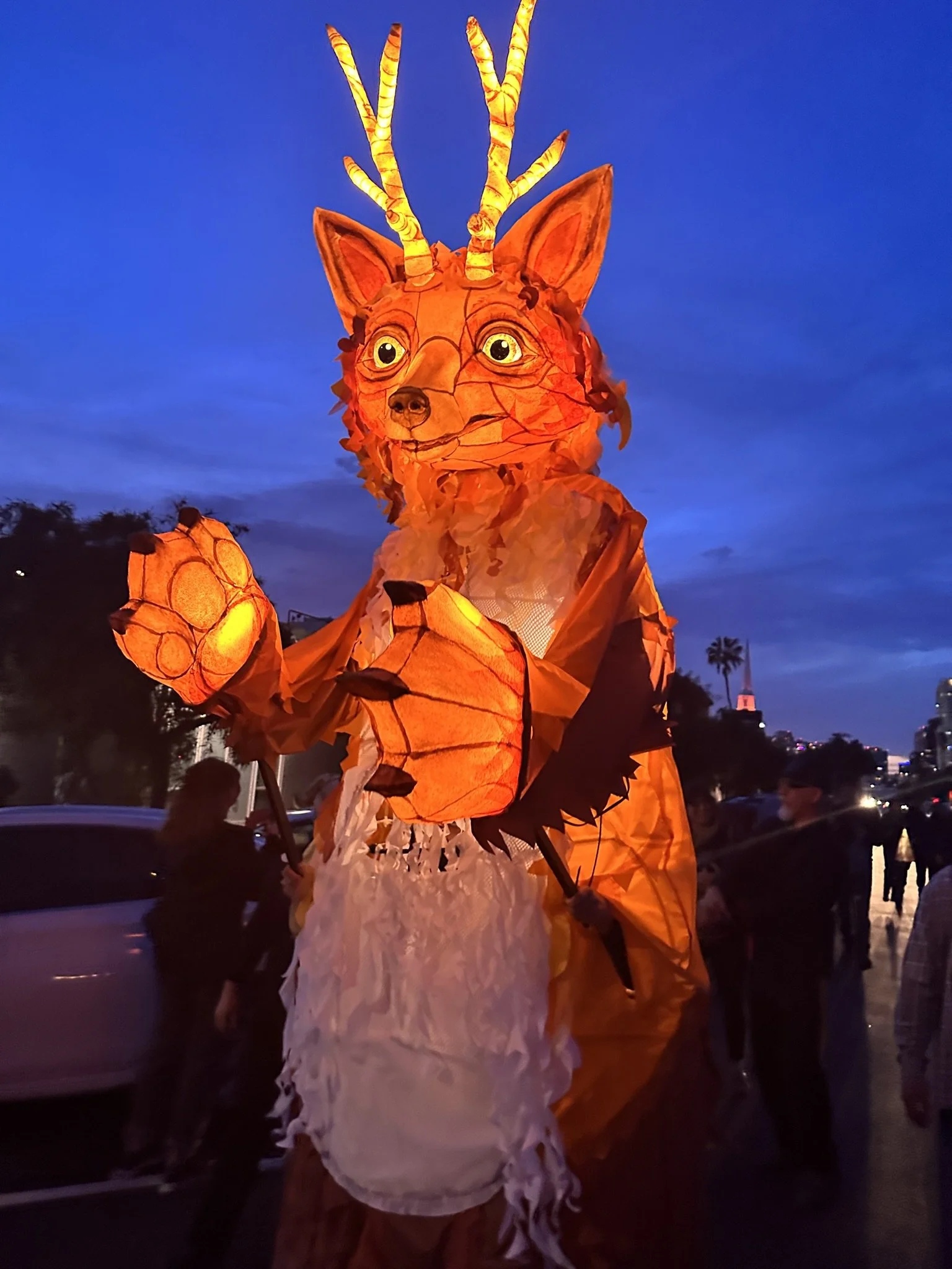 A large illuminated lantern sculpture of a fox with antlers, standing outdoors at dusk.
