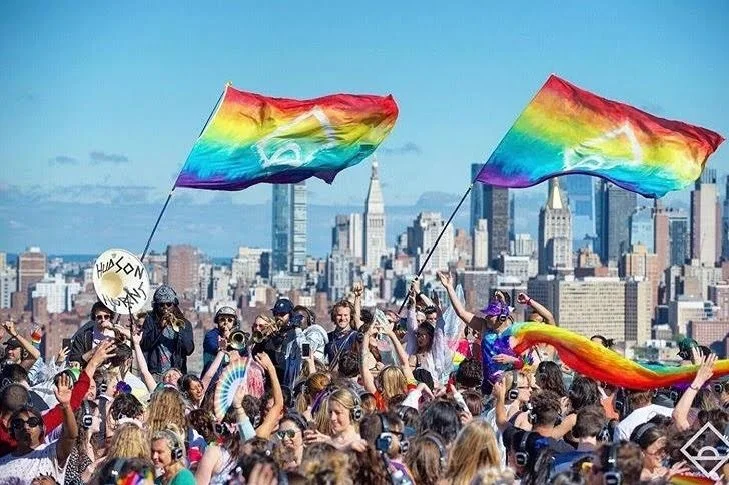 Crowd of people at a pride event holding rainbow flags and banners with city skyline in the background.
