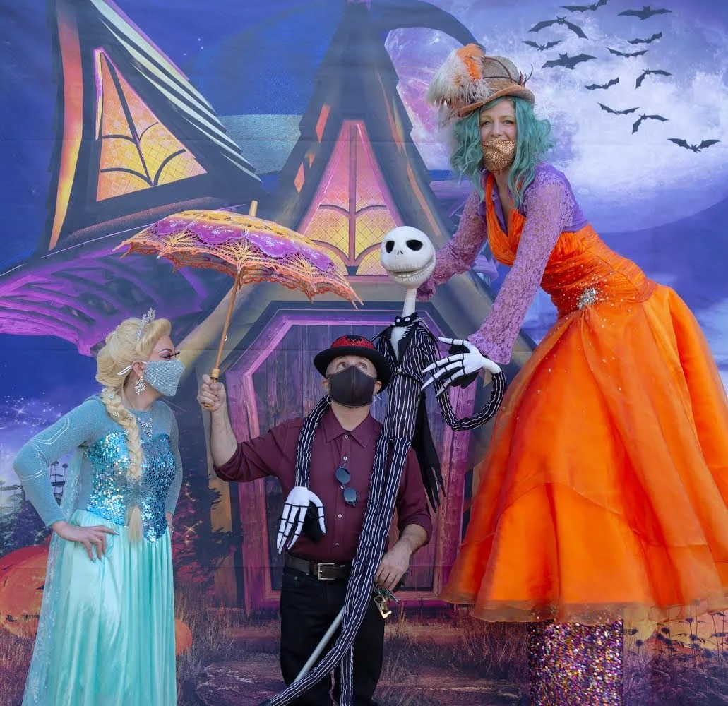 People dressed in Halloween costumes stand in front of a spooky, haunted house backdrop with bats flying in the sky. One person is dressed as Elsa from Frozen, another as a witch, and a third as Jack Skellington from The Nightmare Before Christmas. T