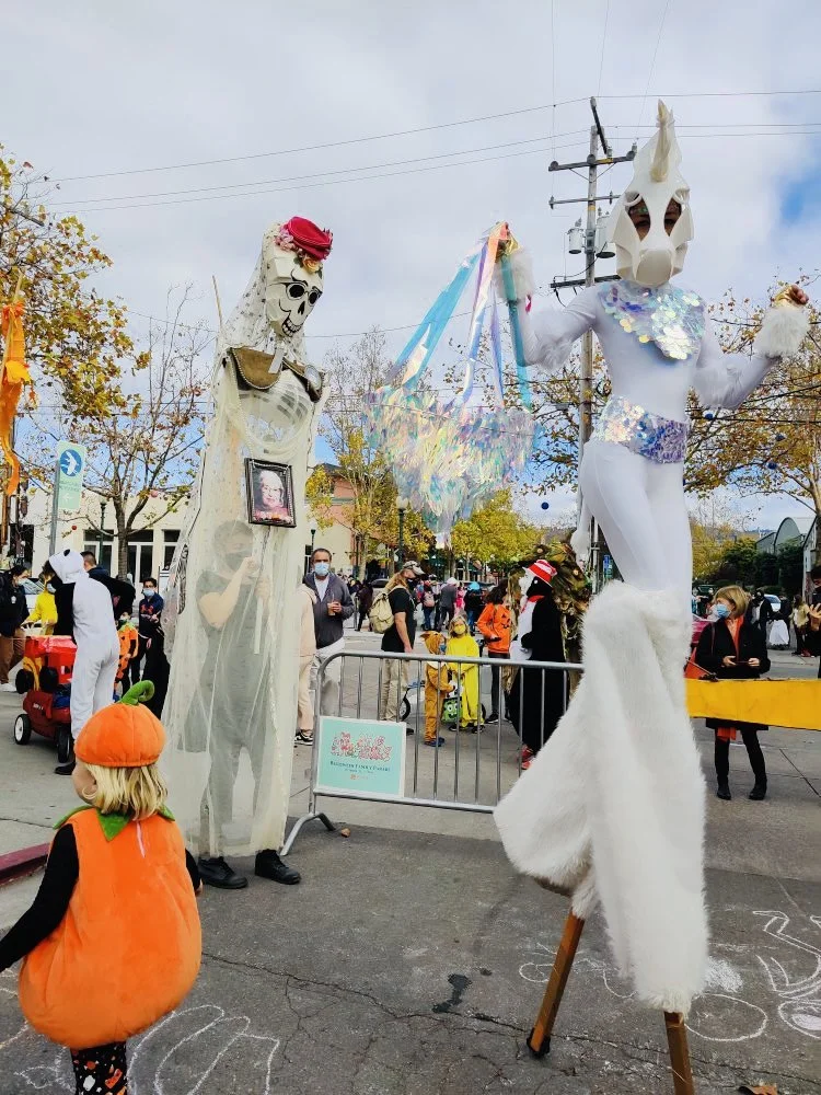 Street scene during a Halloween celebration with large skeleton and unicorn masks, costumes, children in costumes, and decorations.