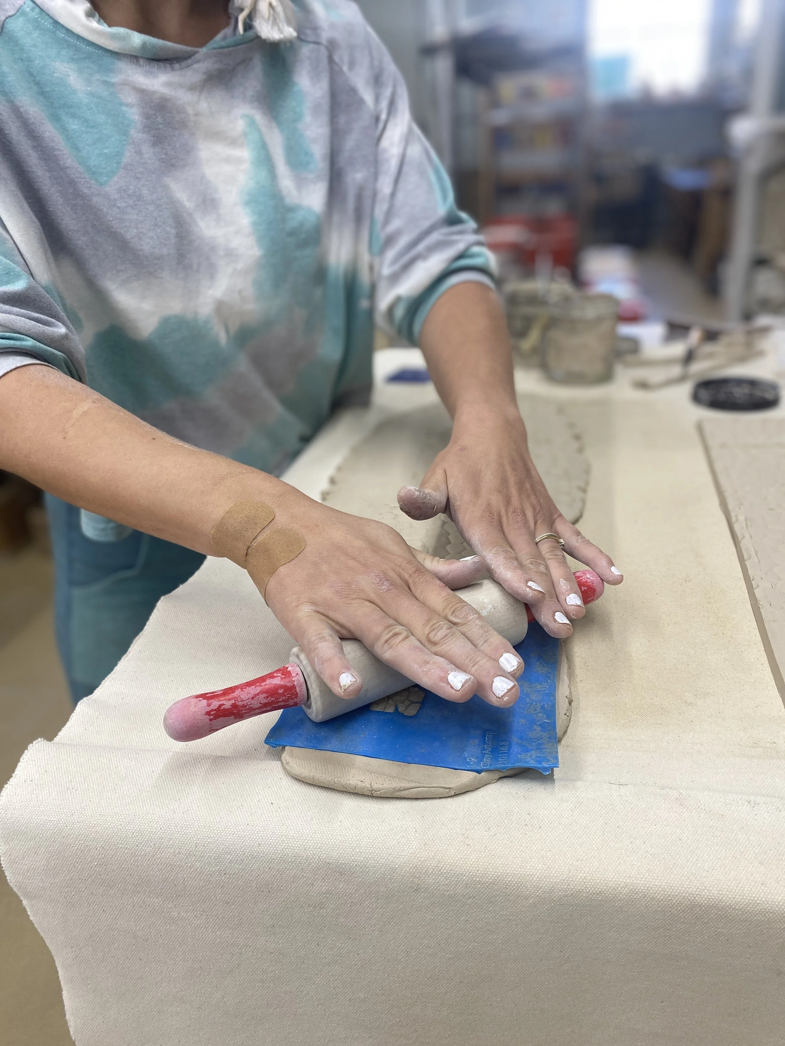 A person using a rolling pin to flatten clay on a work surface. The person is wearing a tie-dye shirt and has a bandage on their right wrist. The workspace background is filled with art supplies and tools.