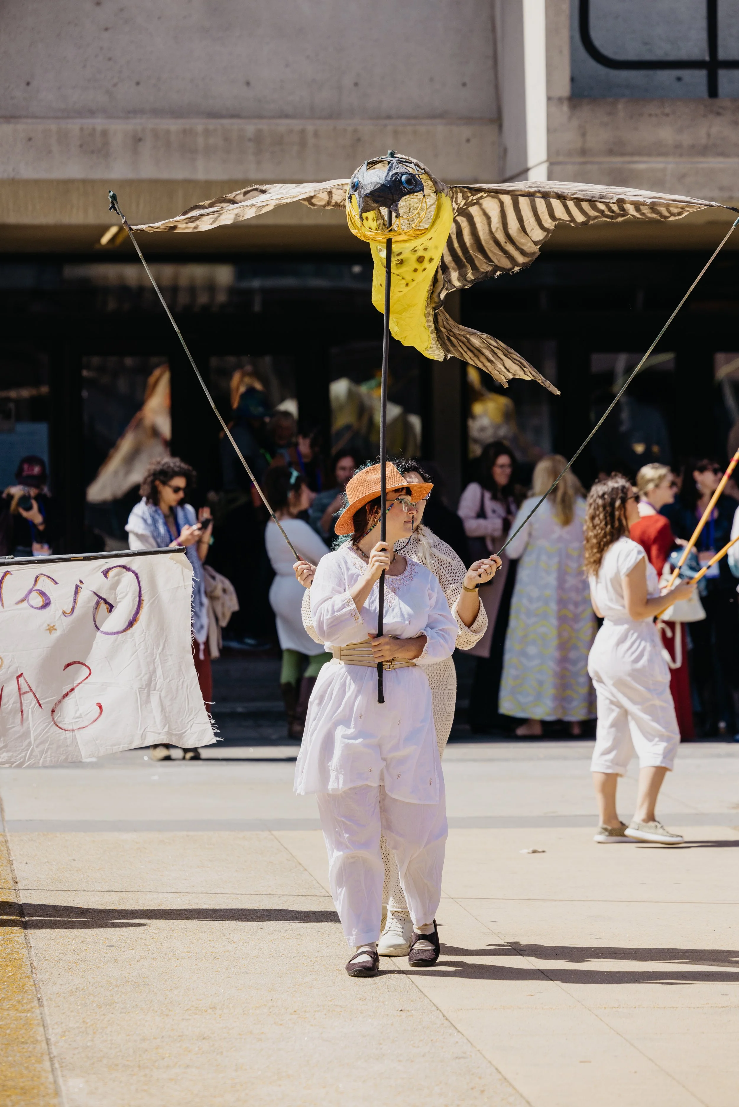 A woman in white clothing, wearing a brown hat and glasses, holding a stick with a large bird-shaped puppet on it at a parade or demonstration on a city sidewalk. There are other people in the background, some also holding signs and puppets.
