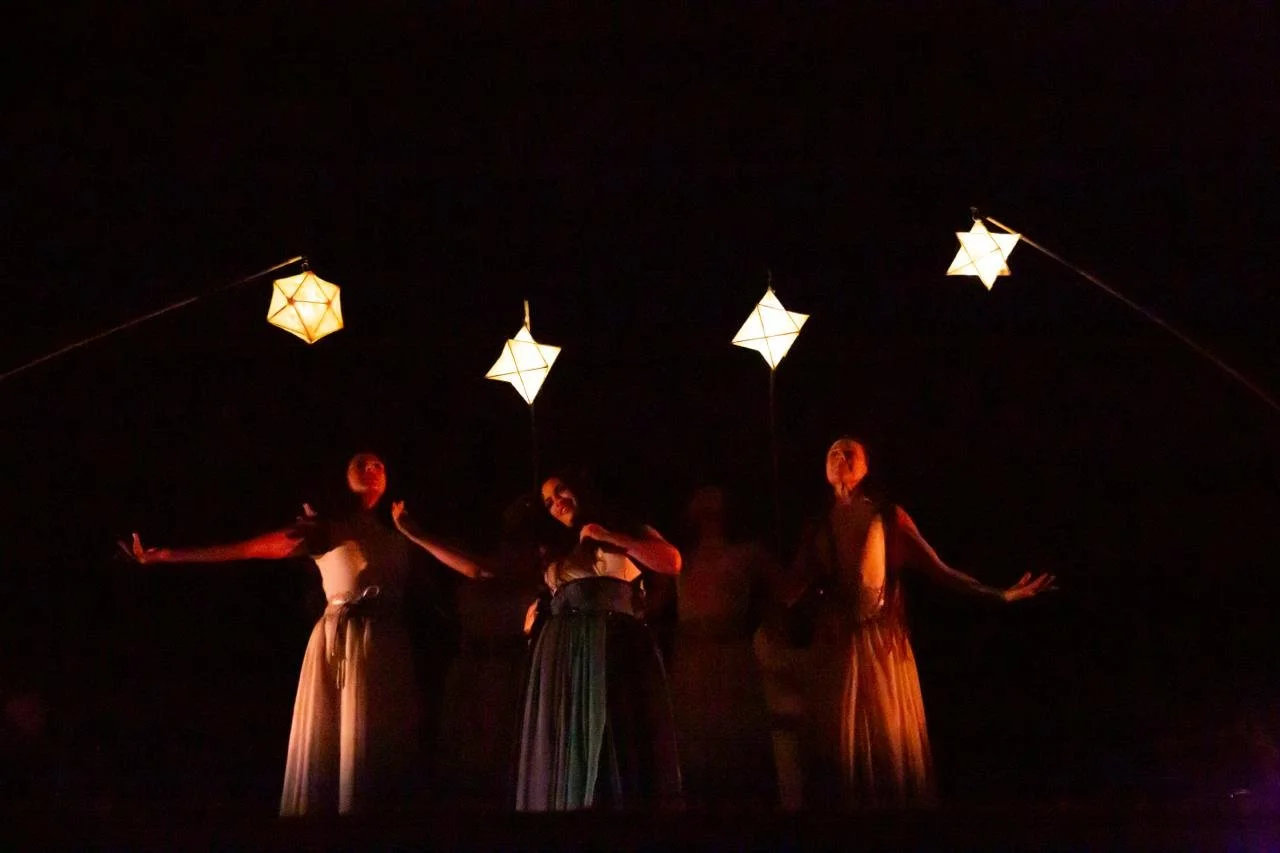 Performance scene with three women in long skirts and dark tops, illuminated by hanging star-shaped lanterns in a dark setting.