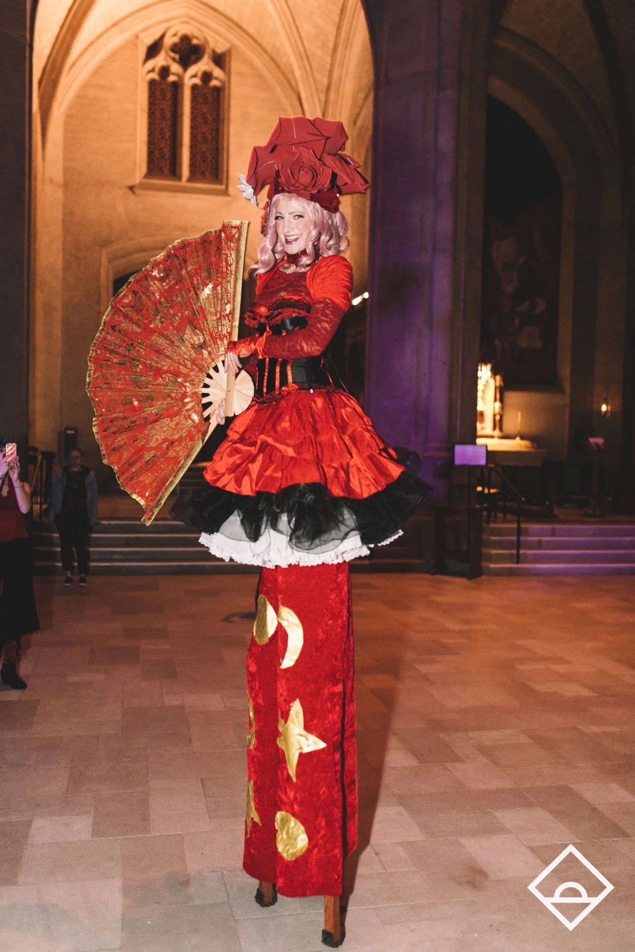 A woman in a red and black costume standing on stilts inside a church or cathedral, holding a red and gold decorated fan and wearing a large red hat with roses.