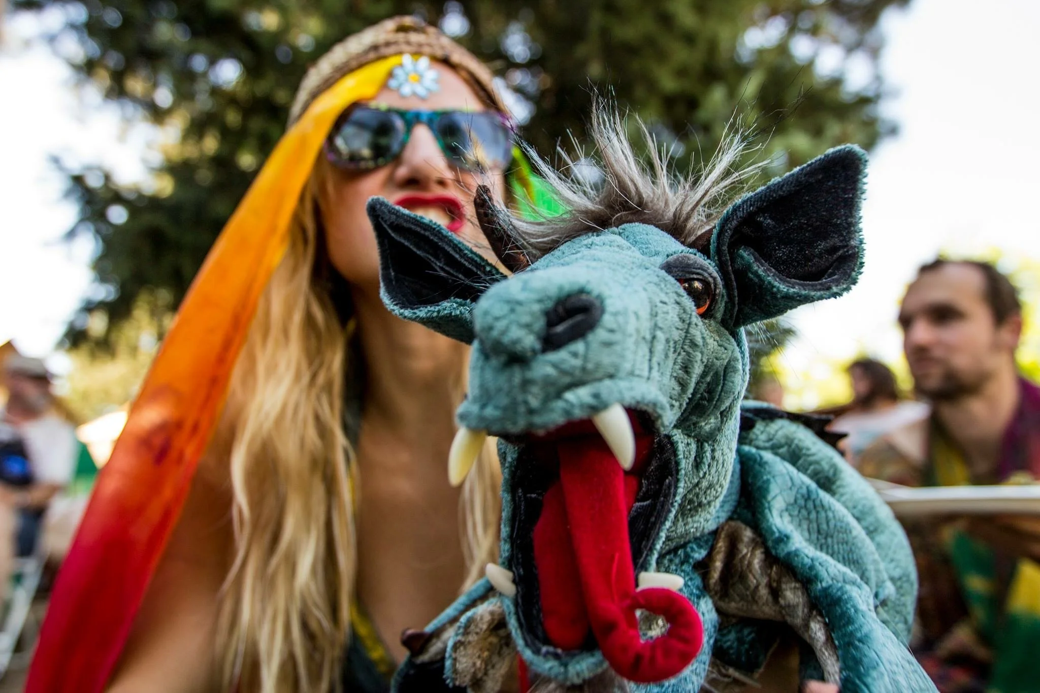 A woman with long blonde hair, wearing sunglasses, a flower headband, and a colorful costume with a red cape, is holding a large plush dragon puppet close to her face. The puppet has a green body, big eyes, and fangs.