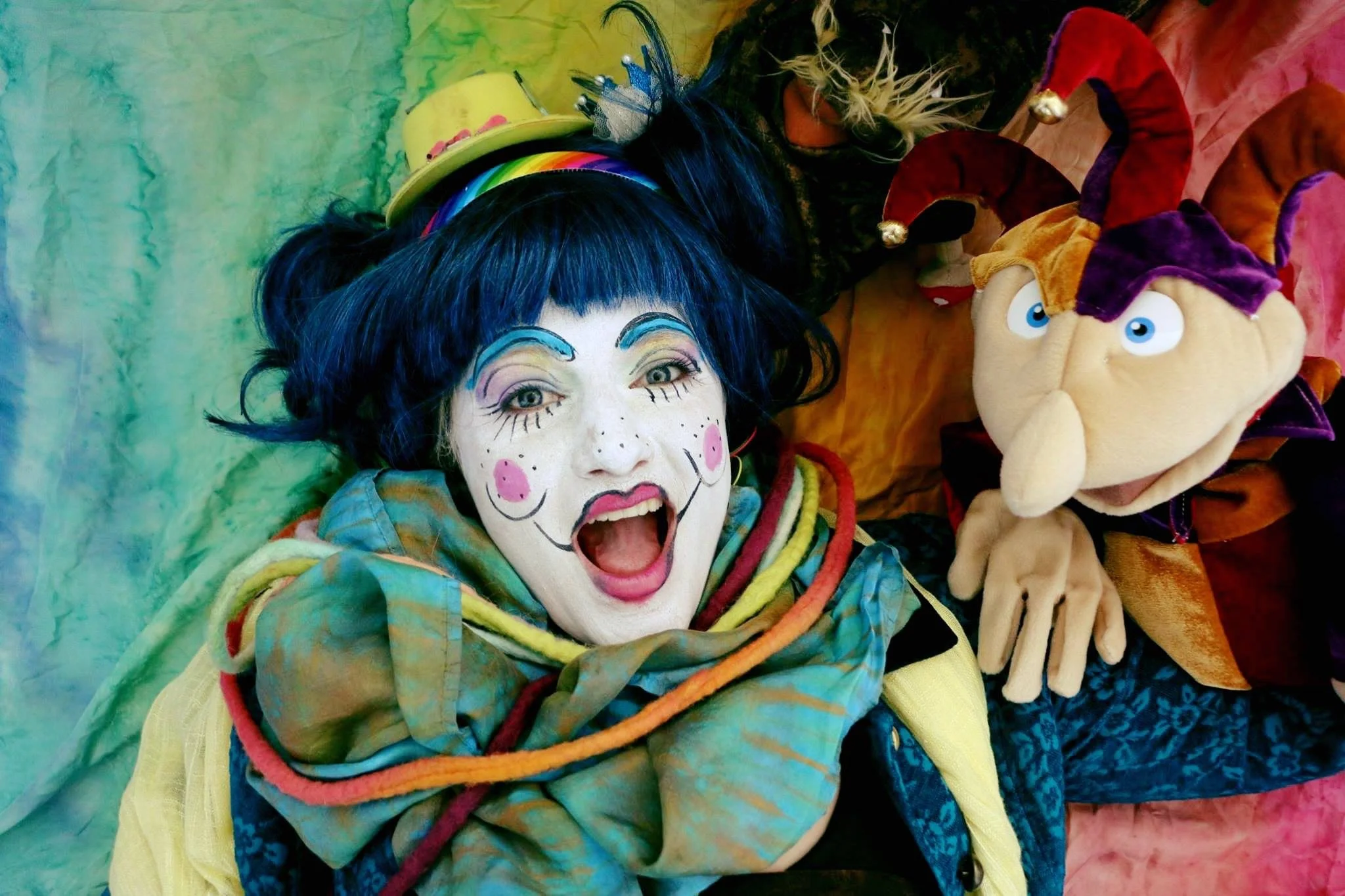 A woman with colorful clown makeup and dark blue hair lying on a multicolored fabric, smiling with an open mouth, next to a large plush toy of a cartoon wizard with a long nose and a red and purple hat.
