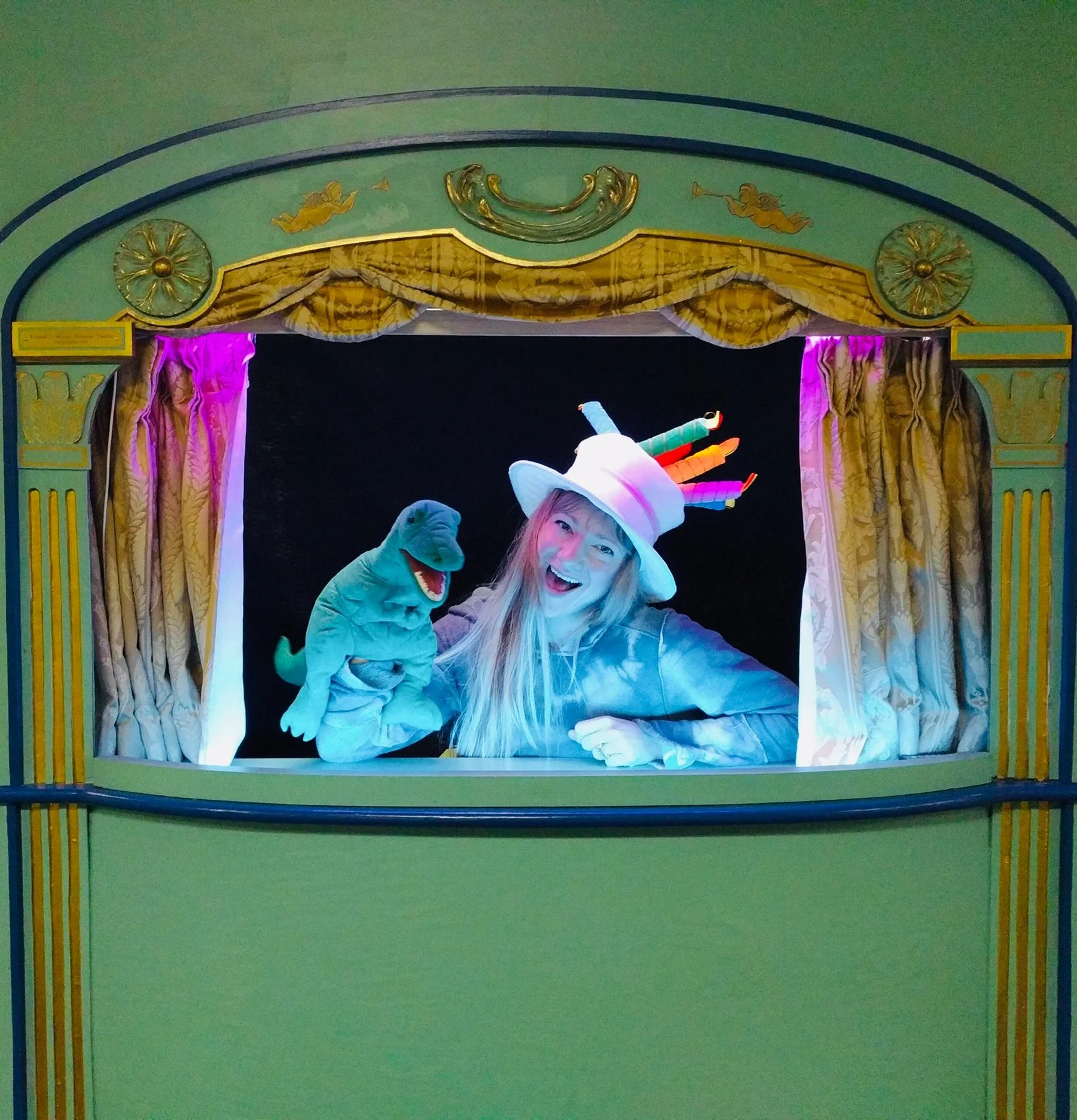 A person dressed as a puppet creator wearing a colorful wizard hat and pink gloves, holding a puppet dinosaur, standing behind a puppet theater stage with gold curtains and green walls.