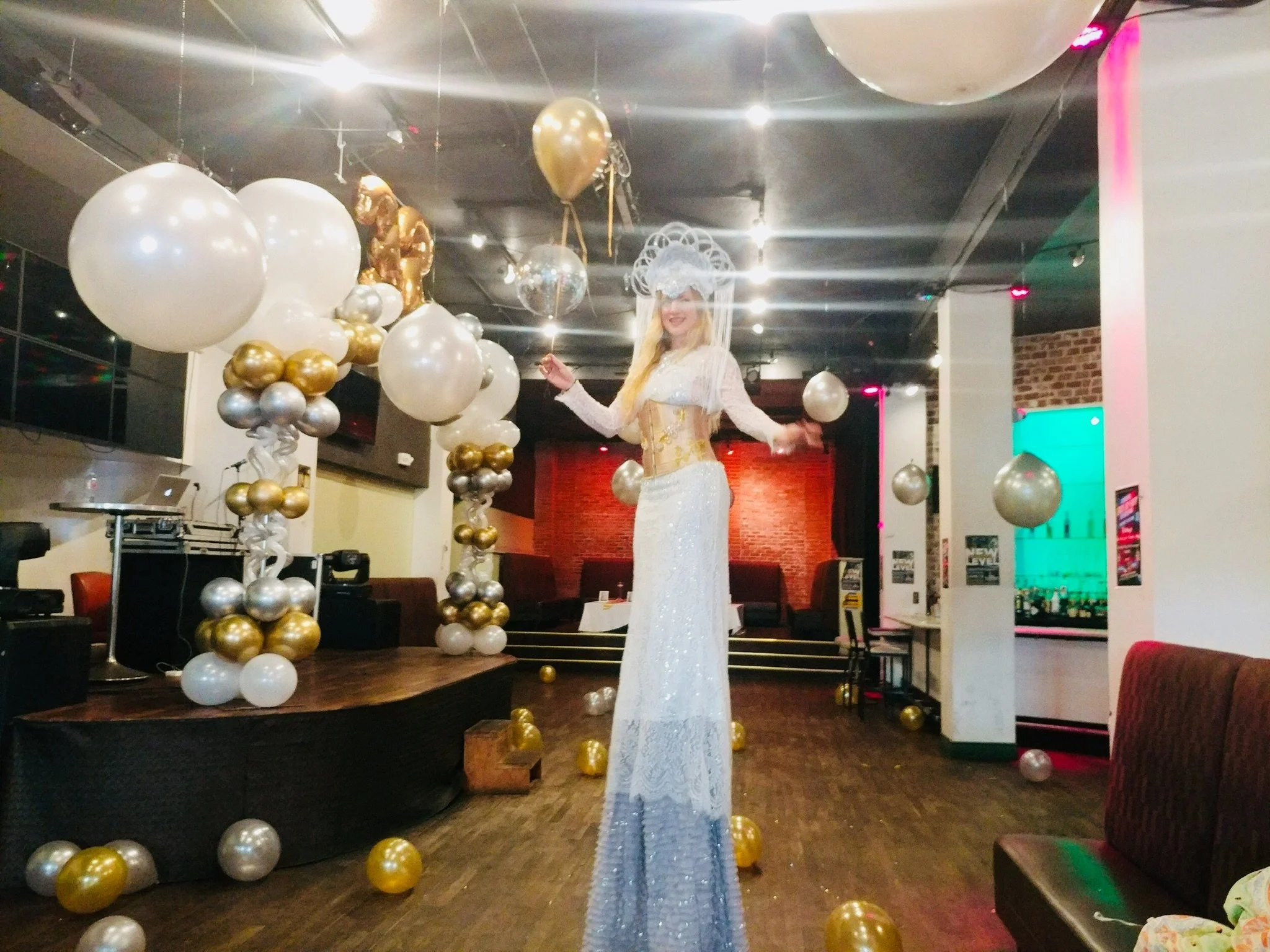 A woman in a white, glittery dress with a large, decorative white headpiece stands on stilts in a decorated venue. The venue features balloons in gold, silver, and white, with some floating from the ceiling and others on the floor. The background inc
