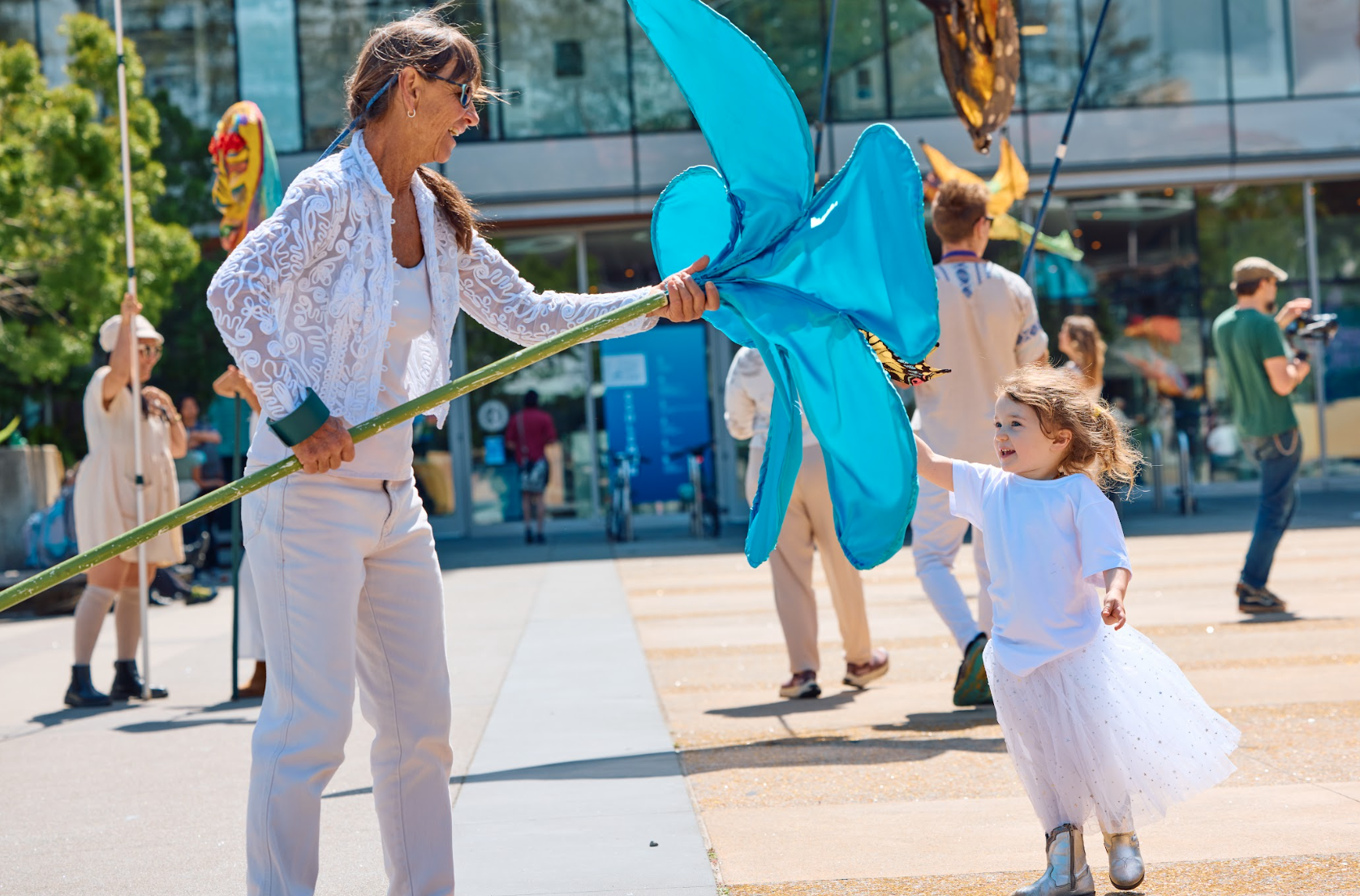 A woman and a young girl enjoying an outdoor event, with the woman holding a large blue butterfly-shaped kite and the girl reaching out to touch it, in front of a modern building with people walking around.