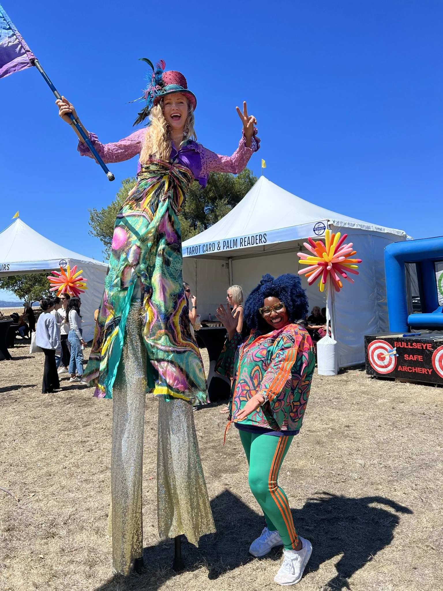 Two colorful women at an outdoor event, one on stilts waving, the other smiling and gesturing. Both are dressed in vibrant, patterned clothing with a carnival-like atmosphere. In the background, there are tents and other people.