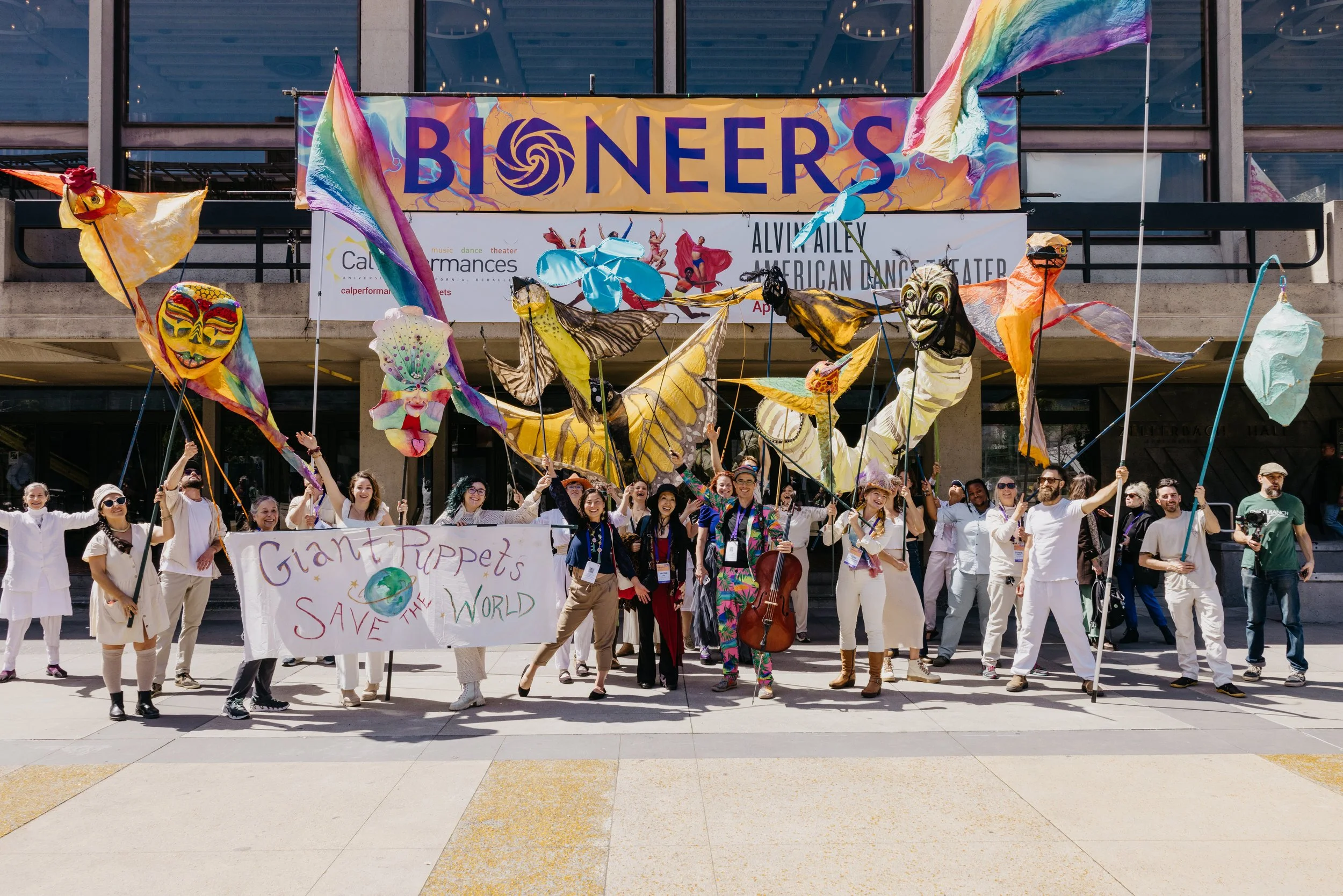 Group of people holding colorful puppets and a banner that reads "Giant Puppets Save the World" outside a building with a large "BIONERS" sign.