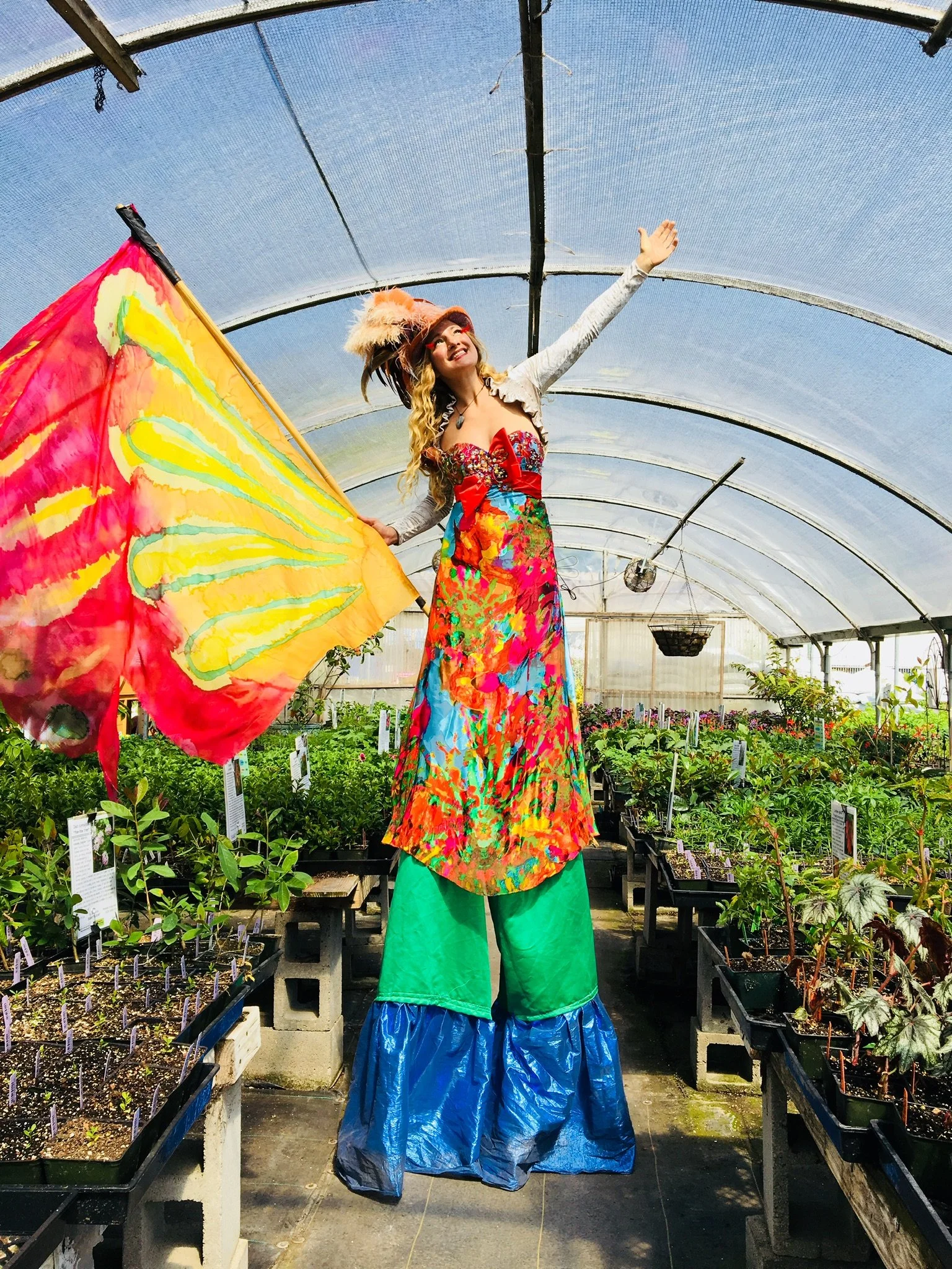 A woman dressed in colorful clothing and a feathered hat, standing on stilts inside a greenhouse, holding a vibrant yellow and red flag with abstract patterns, surrounded by potted plants.