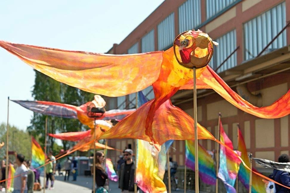 Colorful dragon and bird kites displayed at a pride event, with rainbow flags and people in the background.