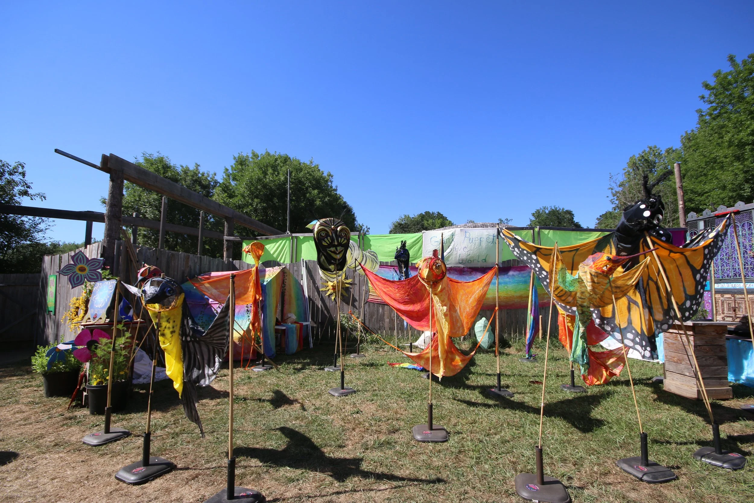 Outdoor display of colorful butterfly and insect-shaped kites with a rainbow backdrop and a wooden fence in the background.