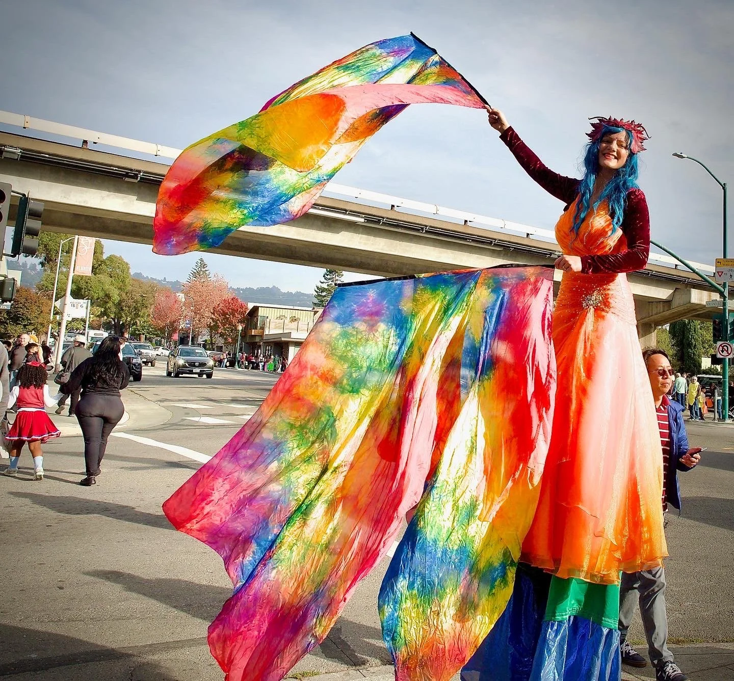 A performer dressed in a colorful clown or carnival costume with a long, orange and pink dress, blue wig, and flower headpiece, standing on stilts and waving rainbow tie-dye flags during a parade.
