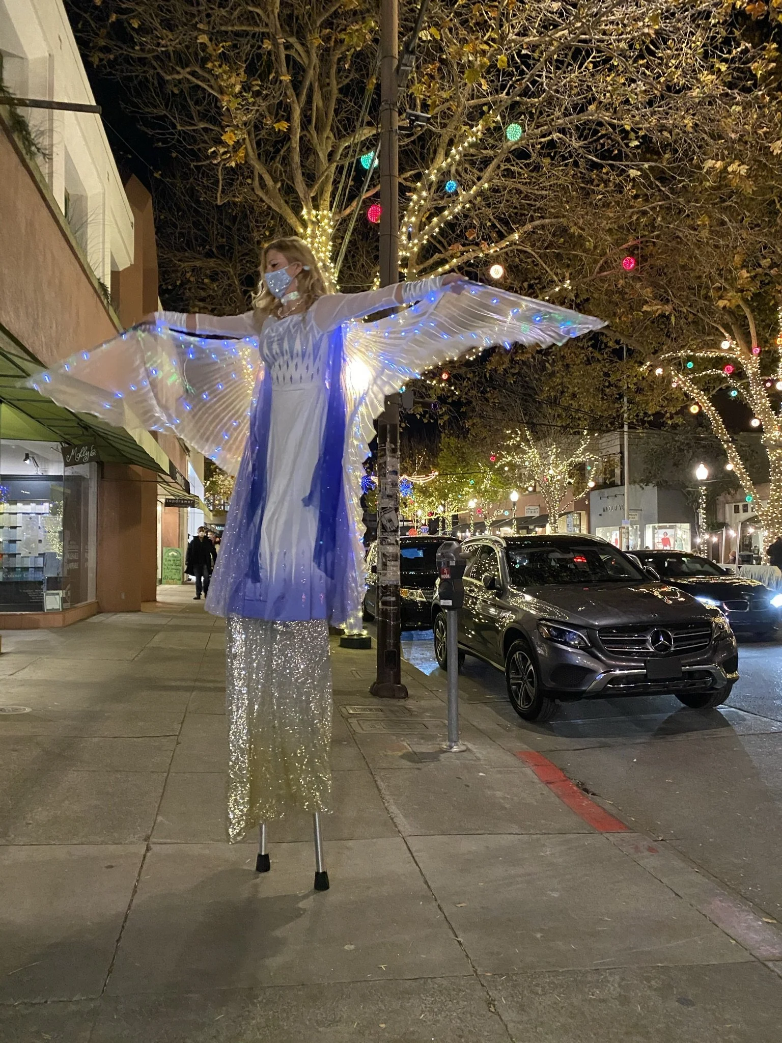 A woman dressed as an angel on stilts with illuminated wings, standing on a city sidewalk at night, surrounded by holiday lights on trees and storefronts.