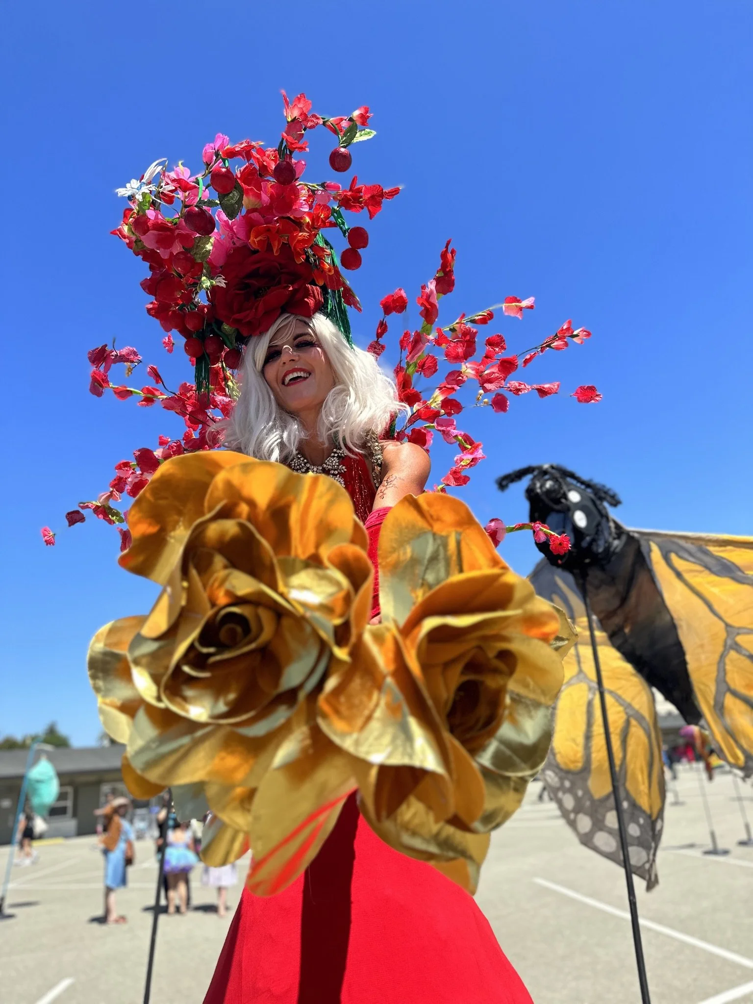 A flamboyant drag queen with long white hair and elaborate floral headpiece, dressed in a red gown, standing outdoors with other people in the background and a clear blue sky.