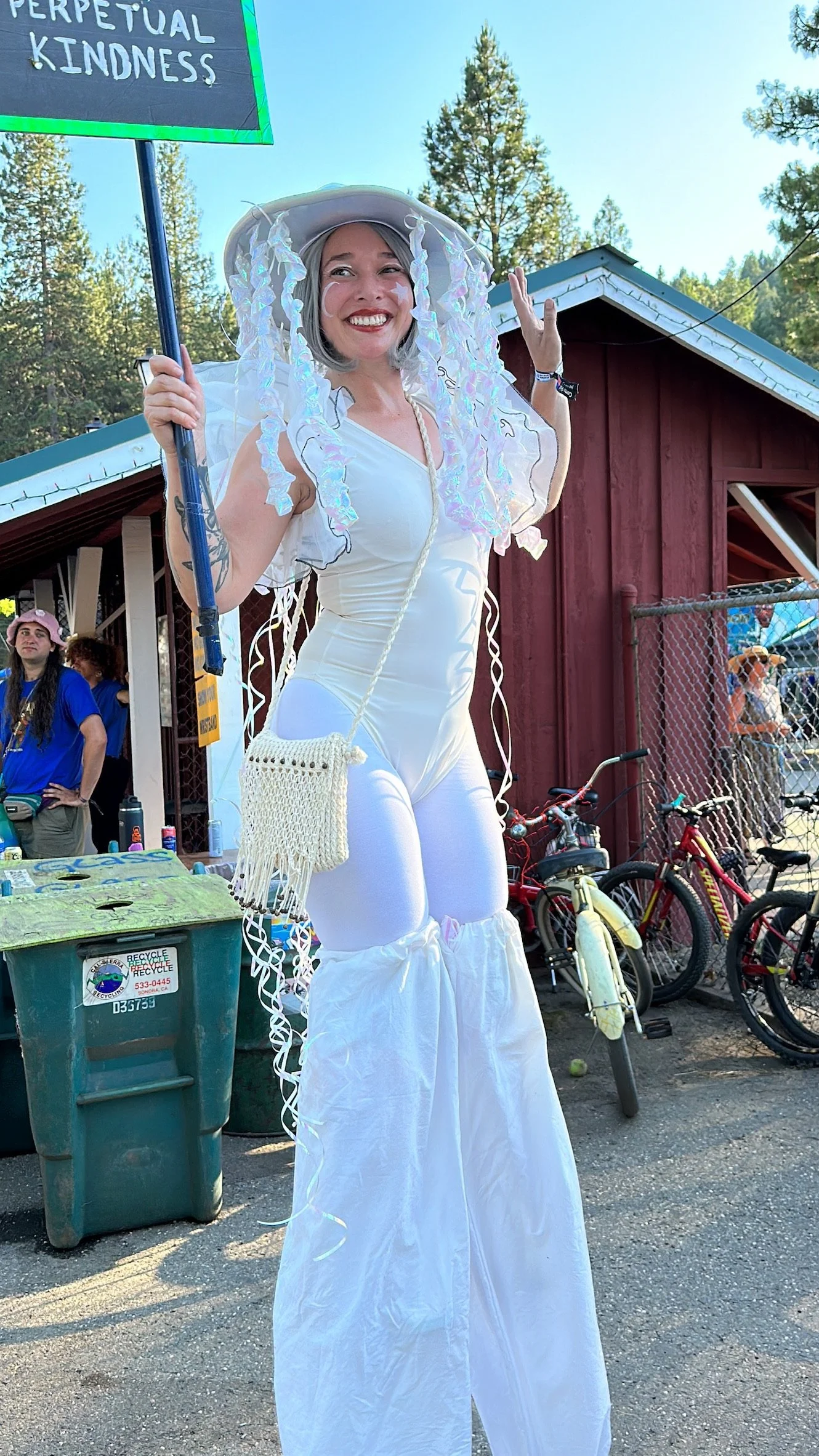 A woman dressed in a white costume with large white pants on stilts, wearing a wide-brimmed hat with iridescent streamers, smiling and holding a sign that says 'Perpetual Kindness'. She is outdoors near a red building, with trees, bicycles, and a few