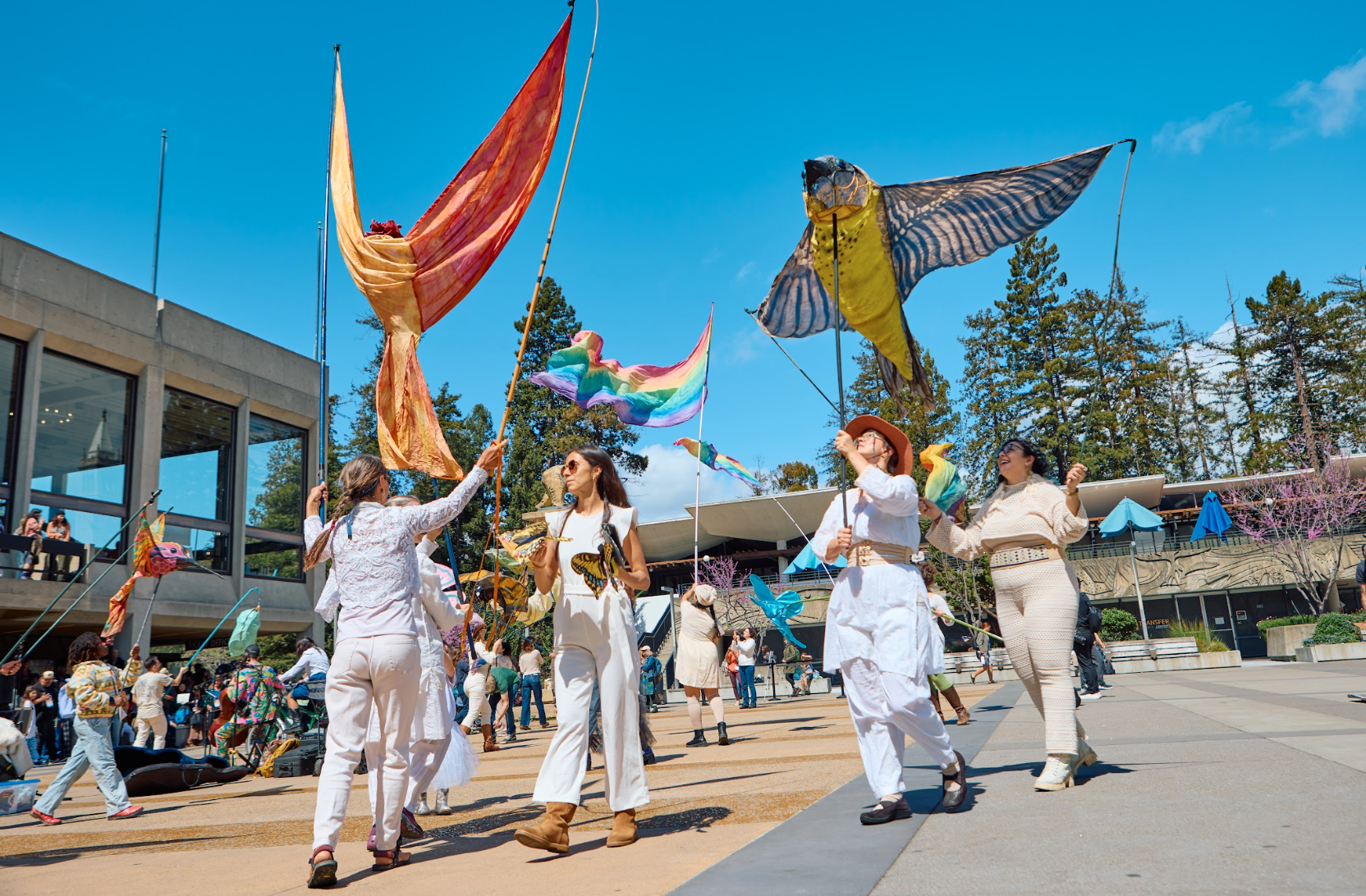 People flying large, colorful kites outdoors on a sunny day with modern buildings and trees in the background.