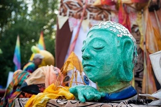 A green ceramic head bust with closed eyes, adorned with small white shells on the top, resting on a surface surrounded by colorful fabrics and objects at an outdoor event.