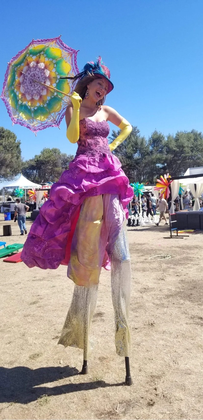 A woman on stilts wearing a colorful pink dress, yellow gloves, and a large colorful hat, holding a vibrant umbrella and smiling at a festival with tents and people in the background.