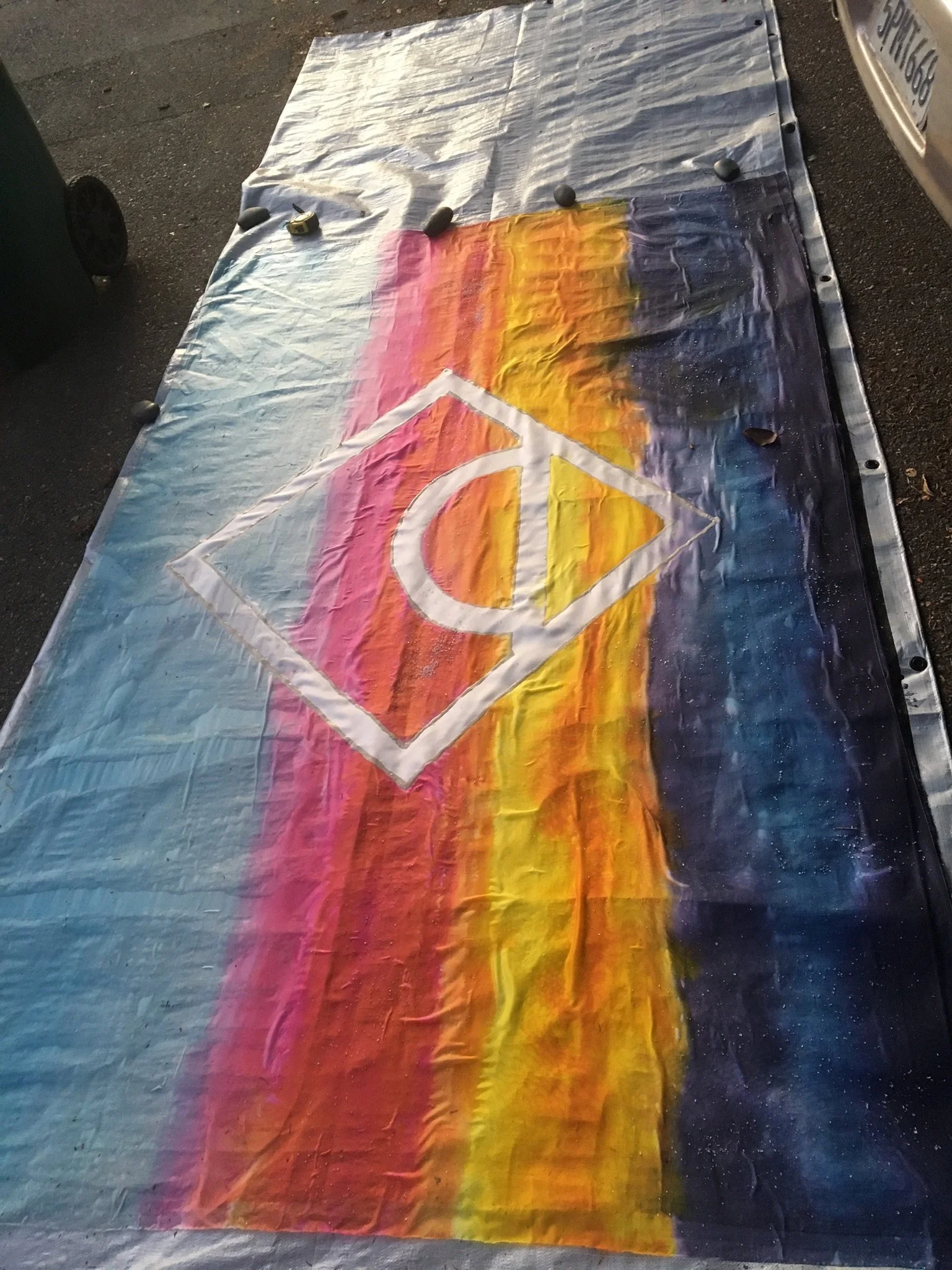 A colorful tie-dye fabric with a peace sign painted in the center, laid on the ground with some rocks around the edges.