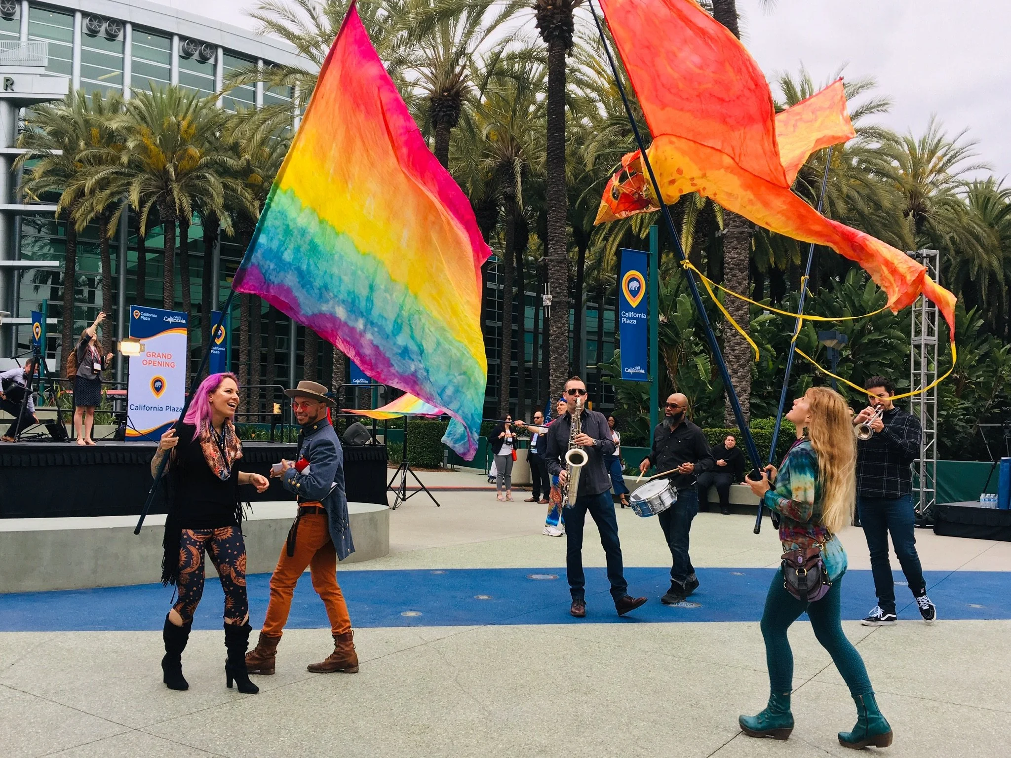 People dancing and playing musical instruments outdoors near a stage at California Plaza, with colorful flags and palm trees in the background.