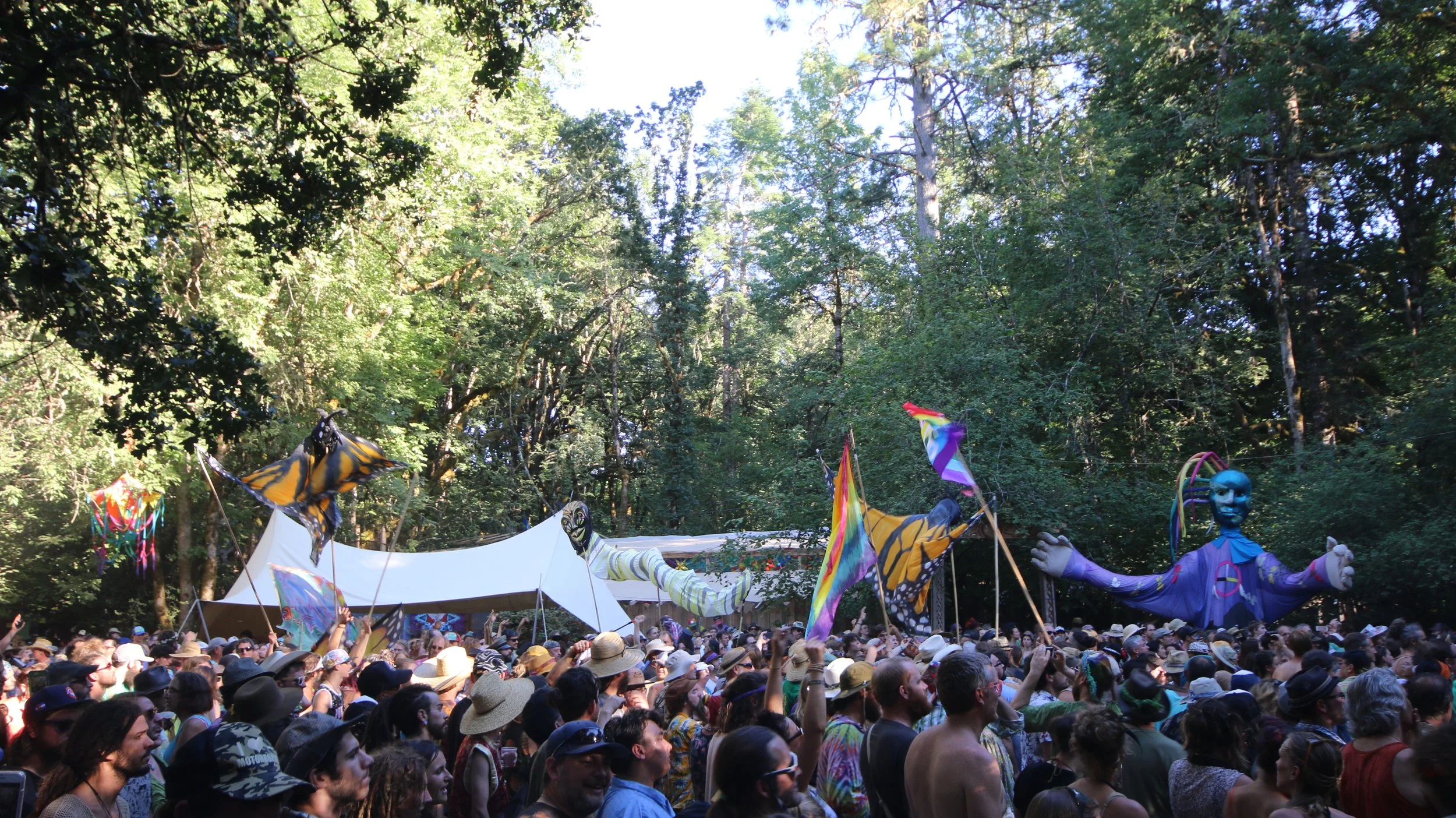 Crowd at outdoor event with colorful flags, puppet-like figures, and a large white canopy, surrounded by trees.