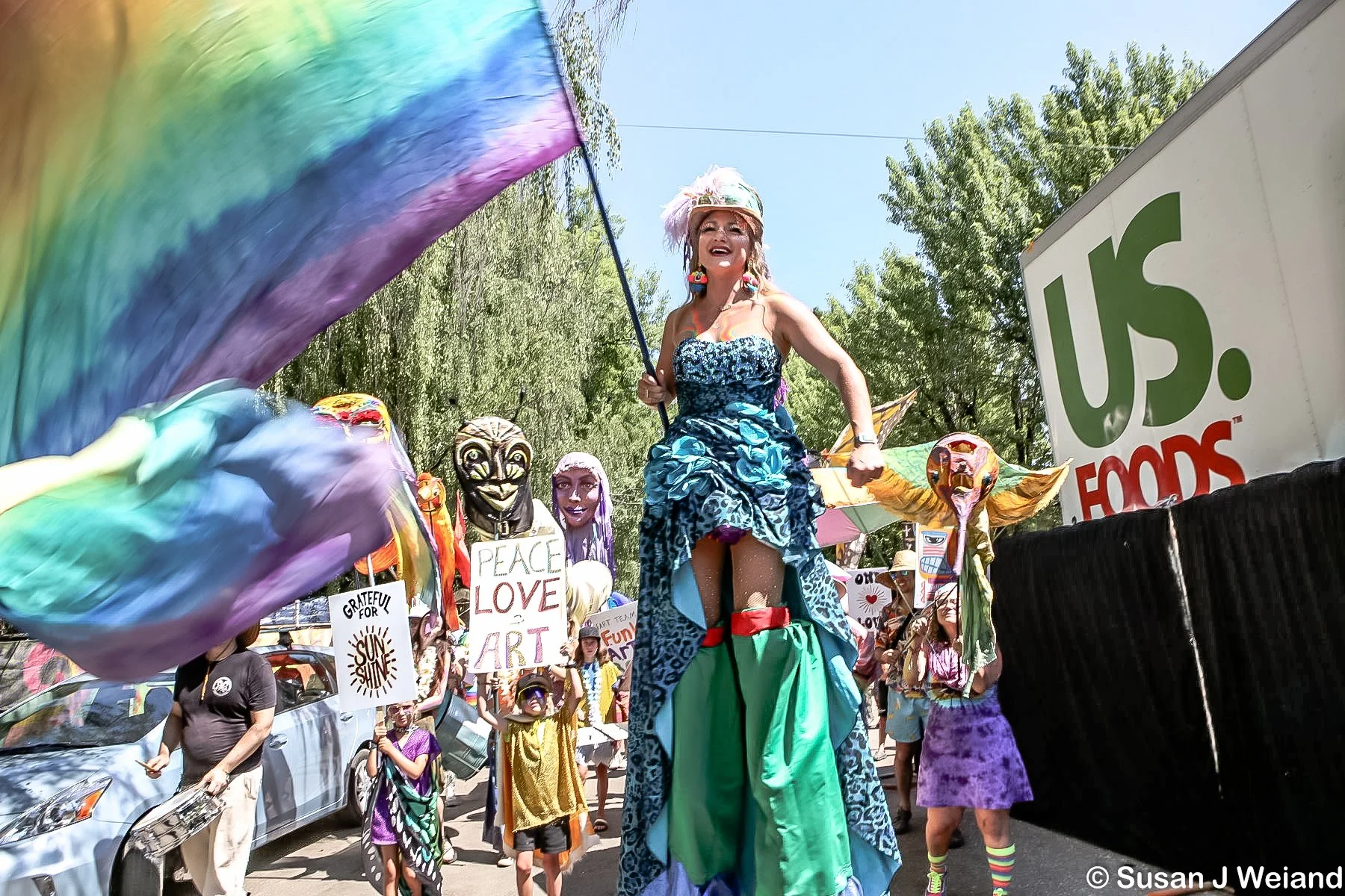 A parade with people carrying signs and puppets, including a woman on stilts wearing a blue dress with a floral pattern, holding a rainbow flag, and wearing a feathered hat. Some signs say 'PEACE LOVE ART' and 'GRATEFUL FOR SUNSHINE.' There are vario