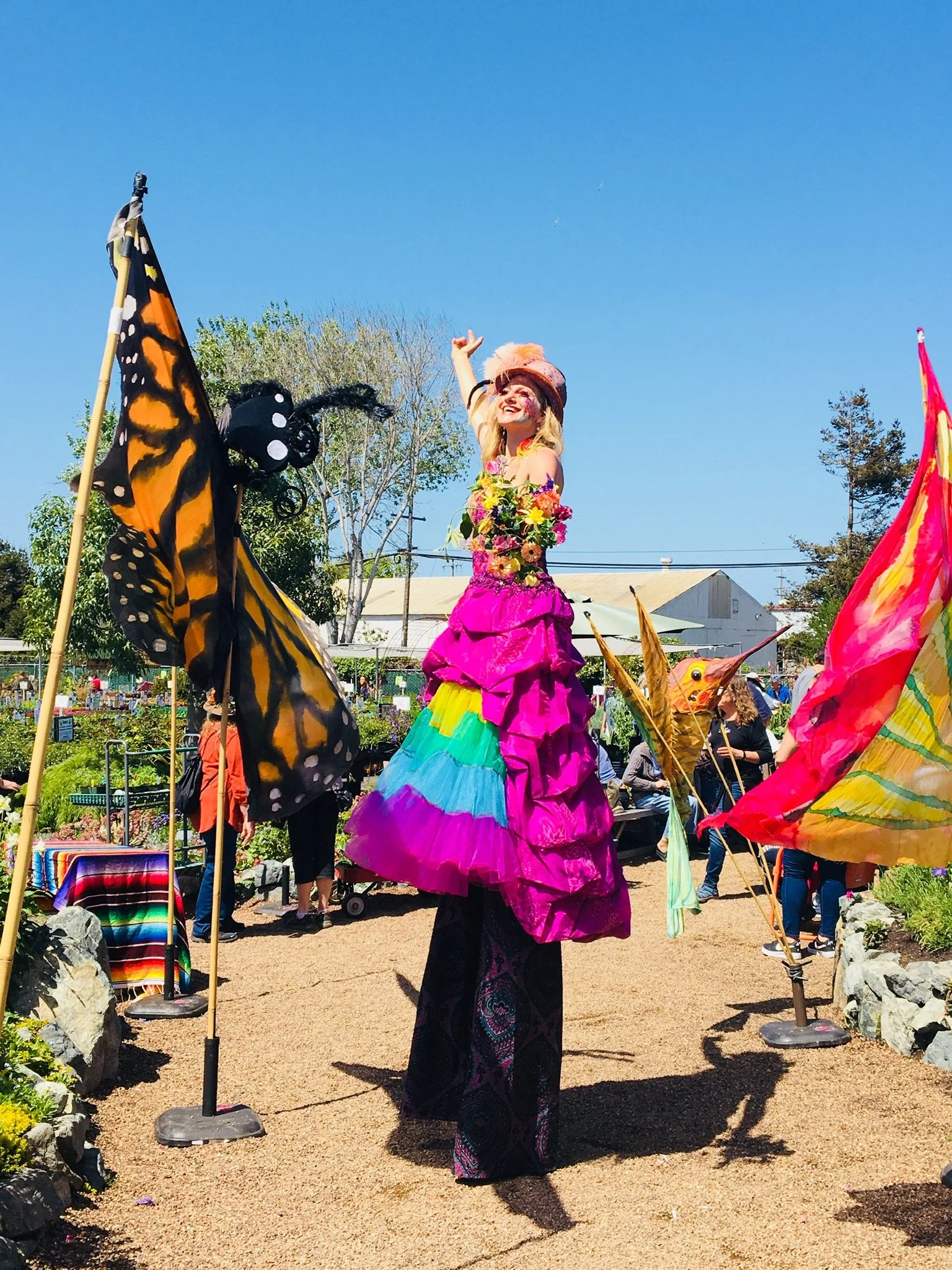 A street performer on stilts dressed in a colorful, layered dress with floral and rainbow patterns, wearing a pink hat with a feather, performing at an outdoor event or festival with flags and people in the background.