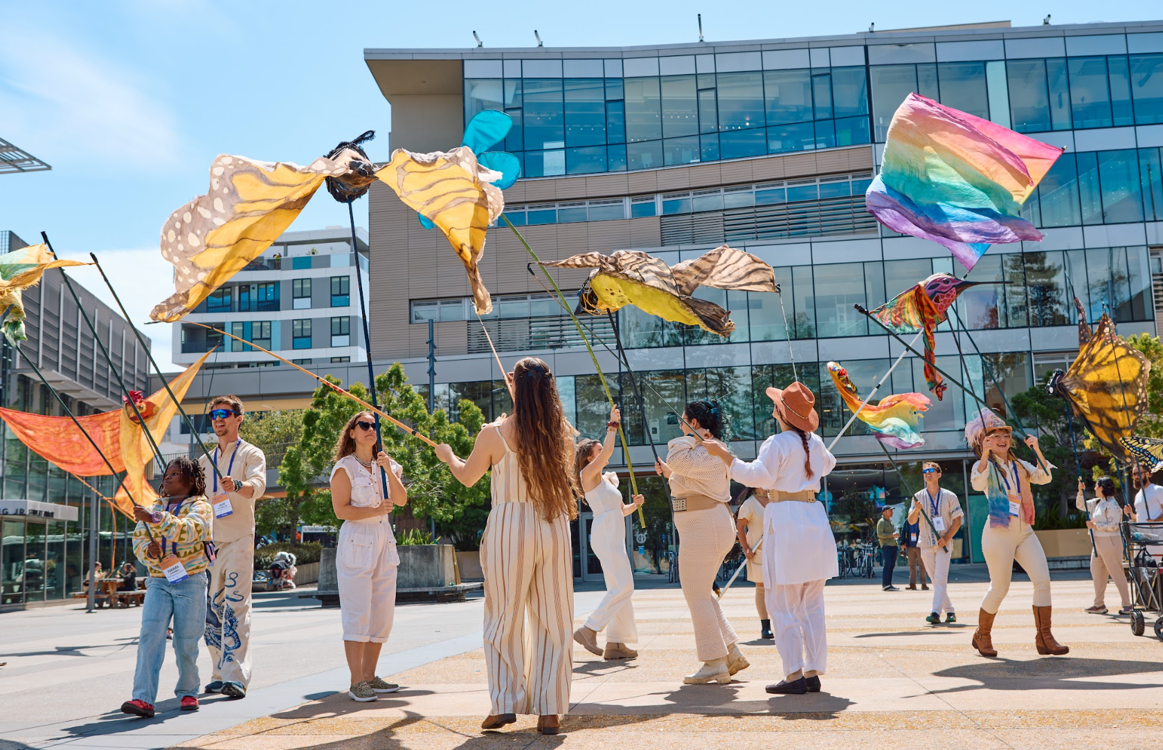 People participating in a kite festival outdoors on a sunny day in an urban setting, with modern glass buildings in the background, and holding colorful, decorated kites.