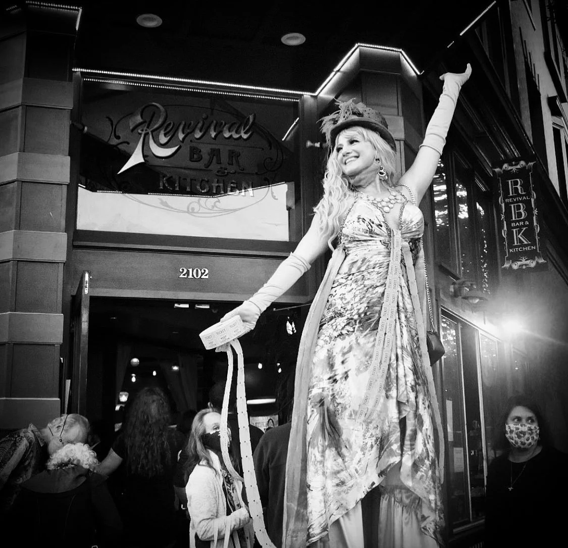 A woman dressed in a vintage, flamboyant outfit, wearing a hat and gloves, stands on a platform outside a bar named 'Revival.' She is smiling and raising her arm, holding tickets, with a group of people, some wearing masks, gathered below.