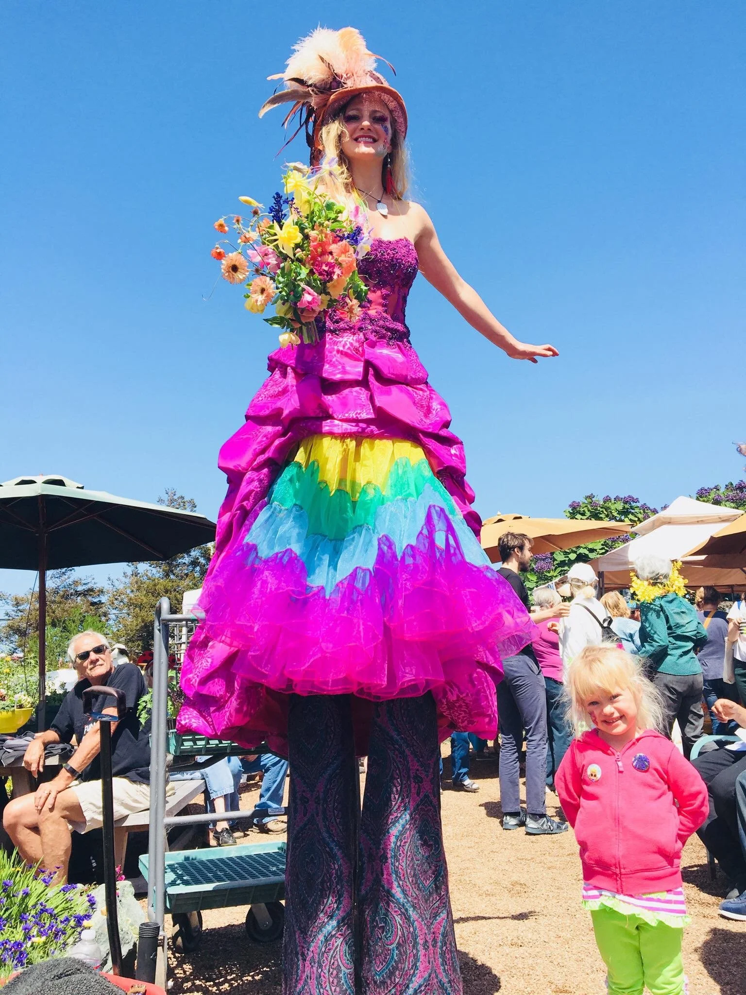 A performer dressed in a colorful, layered dress on stilts, holding a bouquet of flowers, at an outdoor event with people, umbrellas, and blue sky.