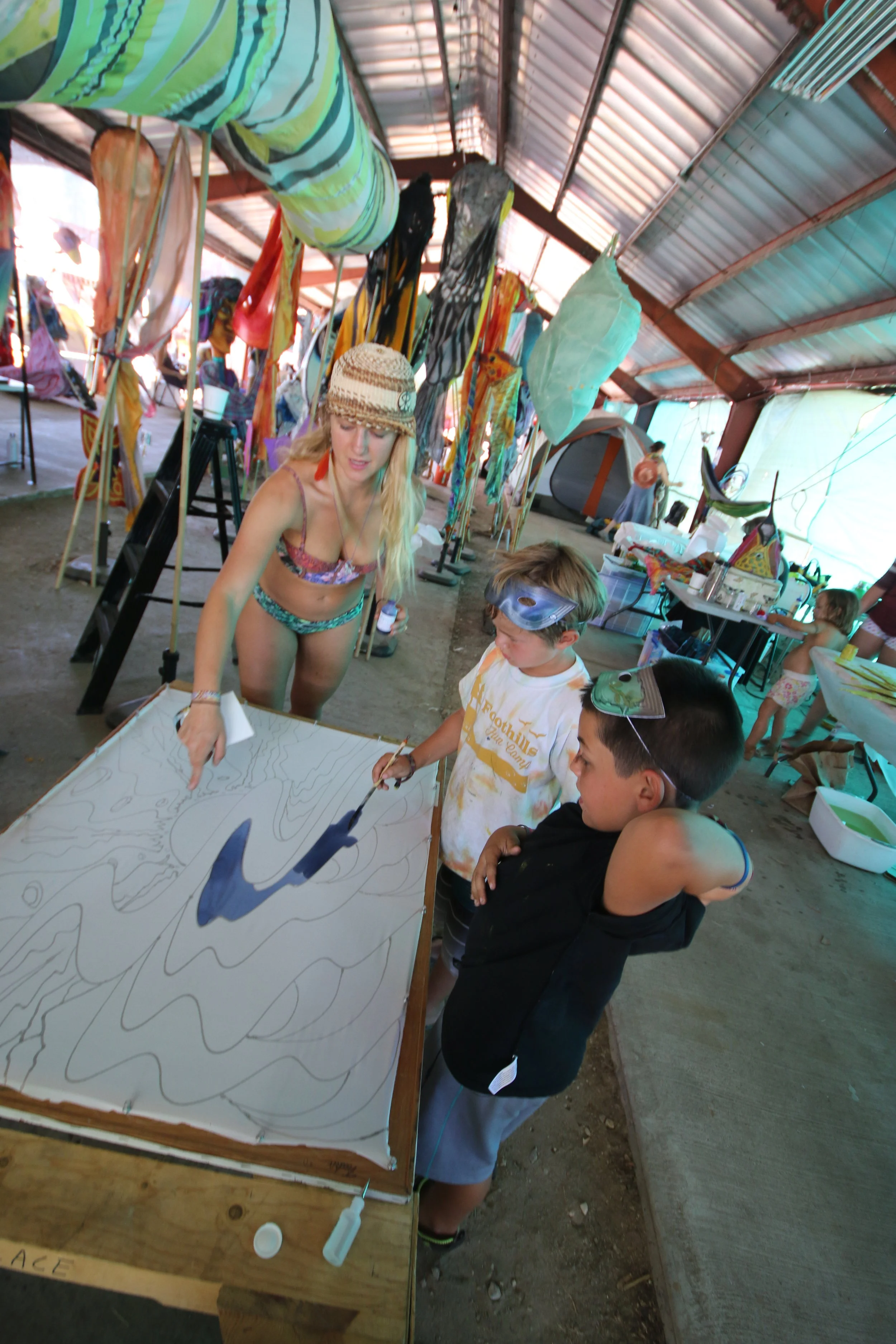 A woman in a bikini and hat demonstrating sand art to two boys with goggles in a beach-themed craft area under a covered pavilion.
