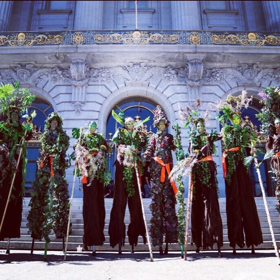 A group of seven people dressed in floral costumes and on stilts, standing on steps in front of a historic building with ornate architectural details and arched windows.