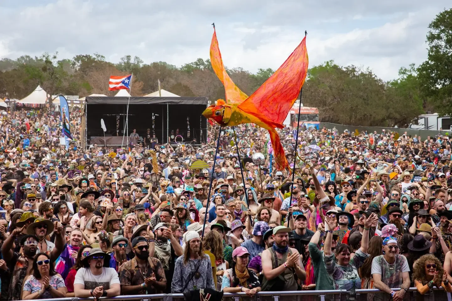 Large crowd attending outdoor music festival with a giant orange and yellow bird puppet in the air.