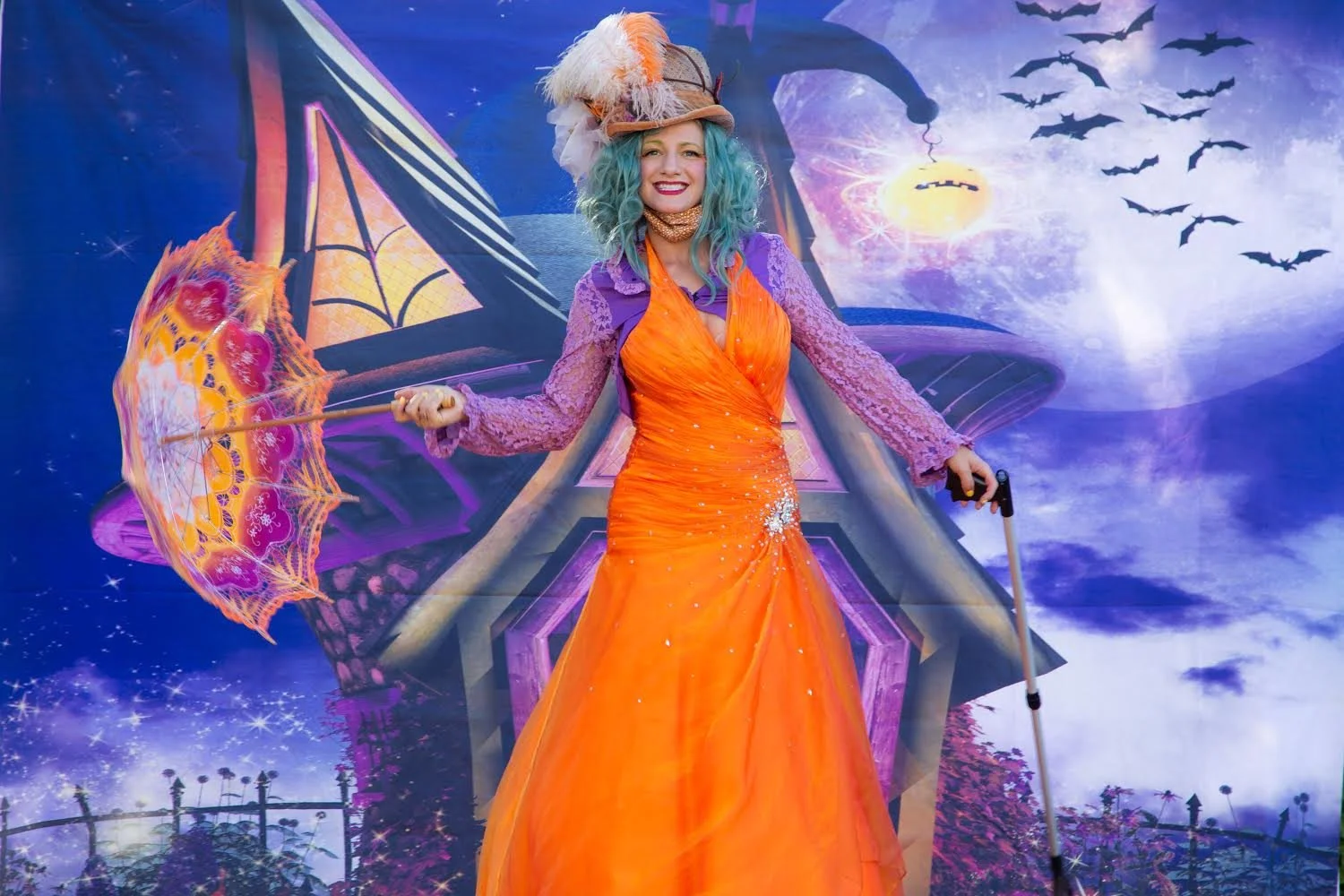 A woman in an orange dress and purple lace sleeves holding an orange umbrella with butterfly and floral designs, standing in front of a colorful fantasy-themed backdrop with a moon, bats, and a house.