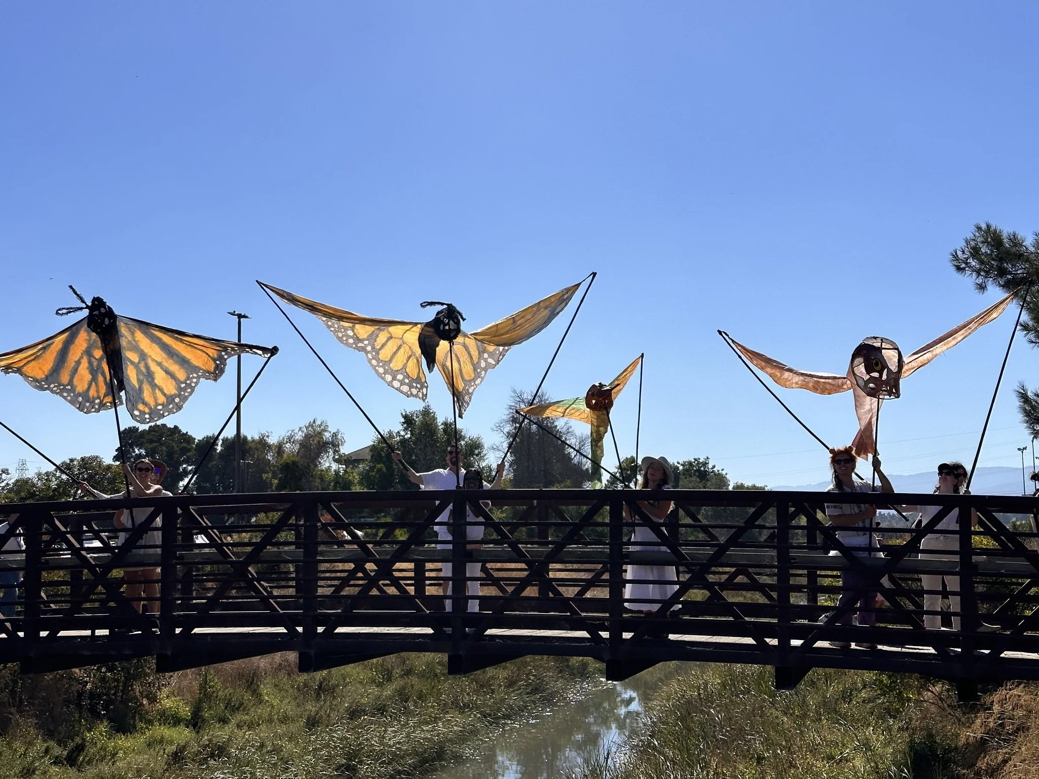 People on a bridge holding large butterfly and skull-shaped kite decorations with a clear blue sky in the background.