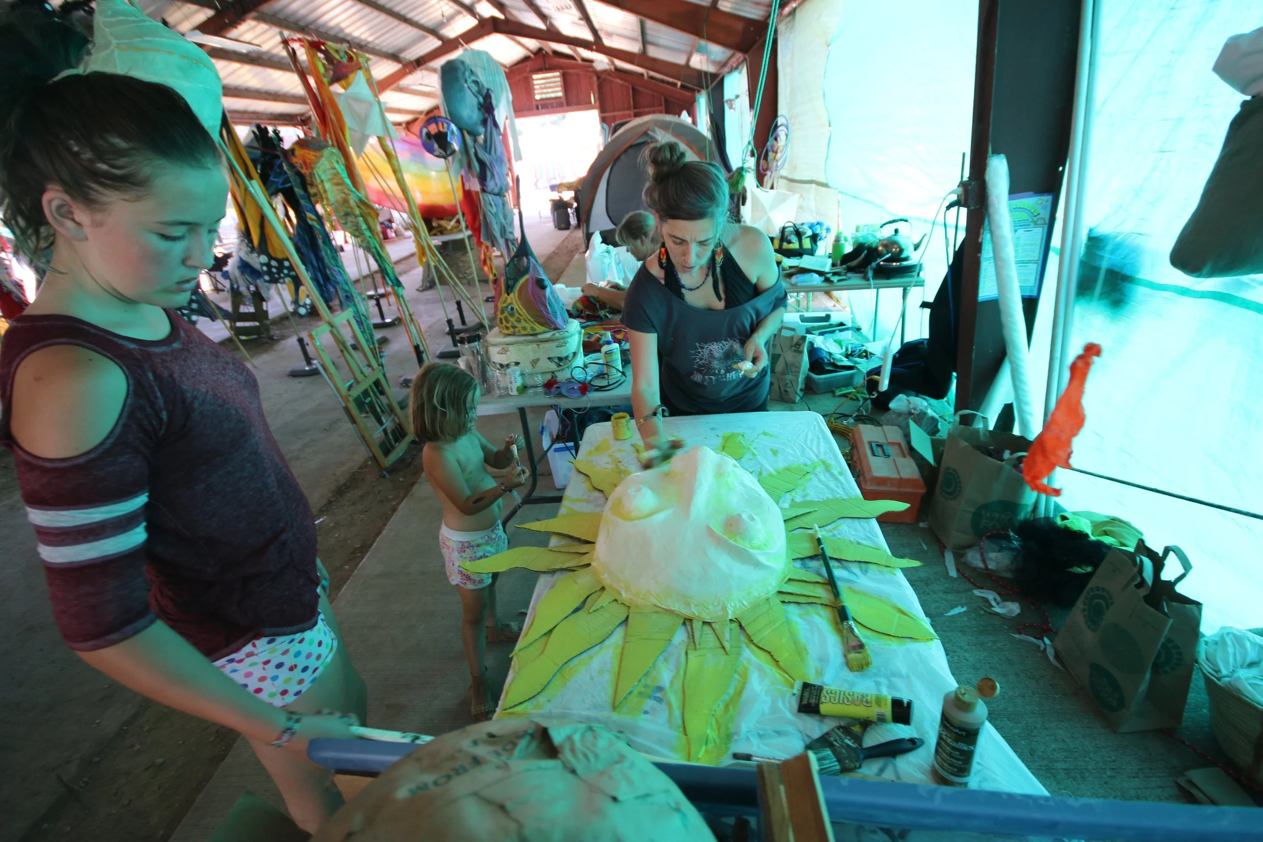 People working on a large sun-themed art project in a spacious, brightly lit workshop or barn, with colorful decorations and art supplies scattered on tables.