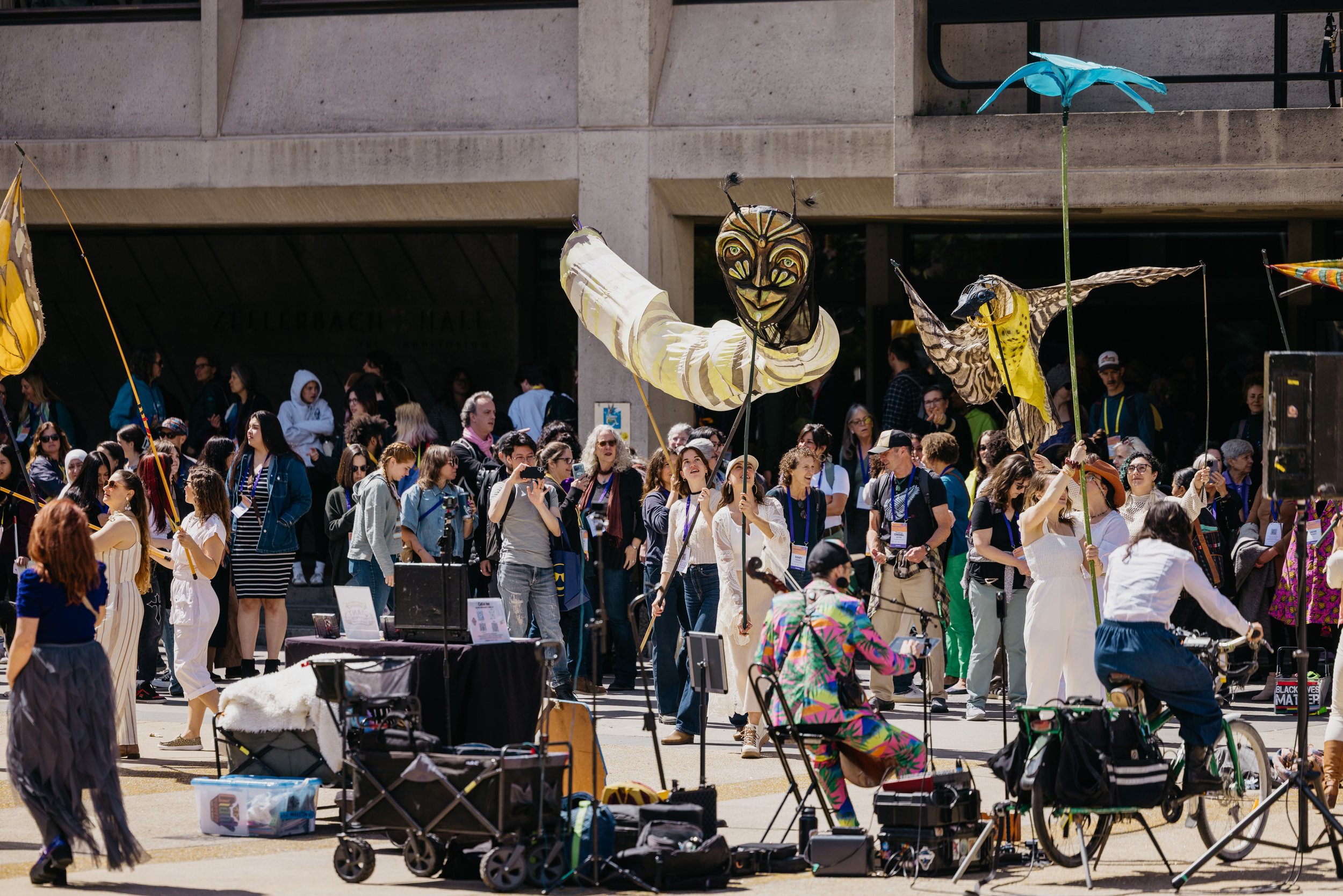 Crowd of people at an outdoor event with large, colorful kite sculptures, including a bee and a caterpillar, held up by participants.