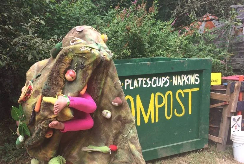 A large, whimsical sculpture of a person climbing a hill made of natural-looking materials, with various objects and fruit attached. In the background, there is a green container with yellow and white text that reads "PLATES CUPS NAPKINS COMPOST."
