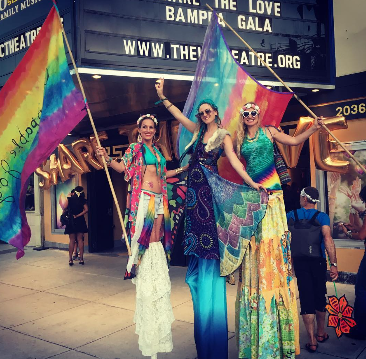 Three women on stilts in colorful, bohemian-style clothing celebrating Pride outside a theater with rainbow flags and gold balloons.