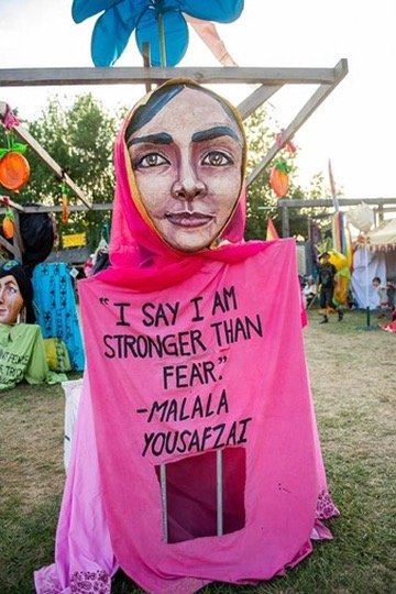 A large puppet with a woman's face and pink headscarf holding a pink sign that says, 'I SAY I AM STRONGER THAN FEAR.' - MALALA YOUSAFZAI, at an outdoor festival.