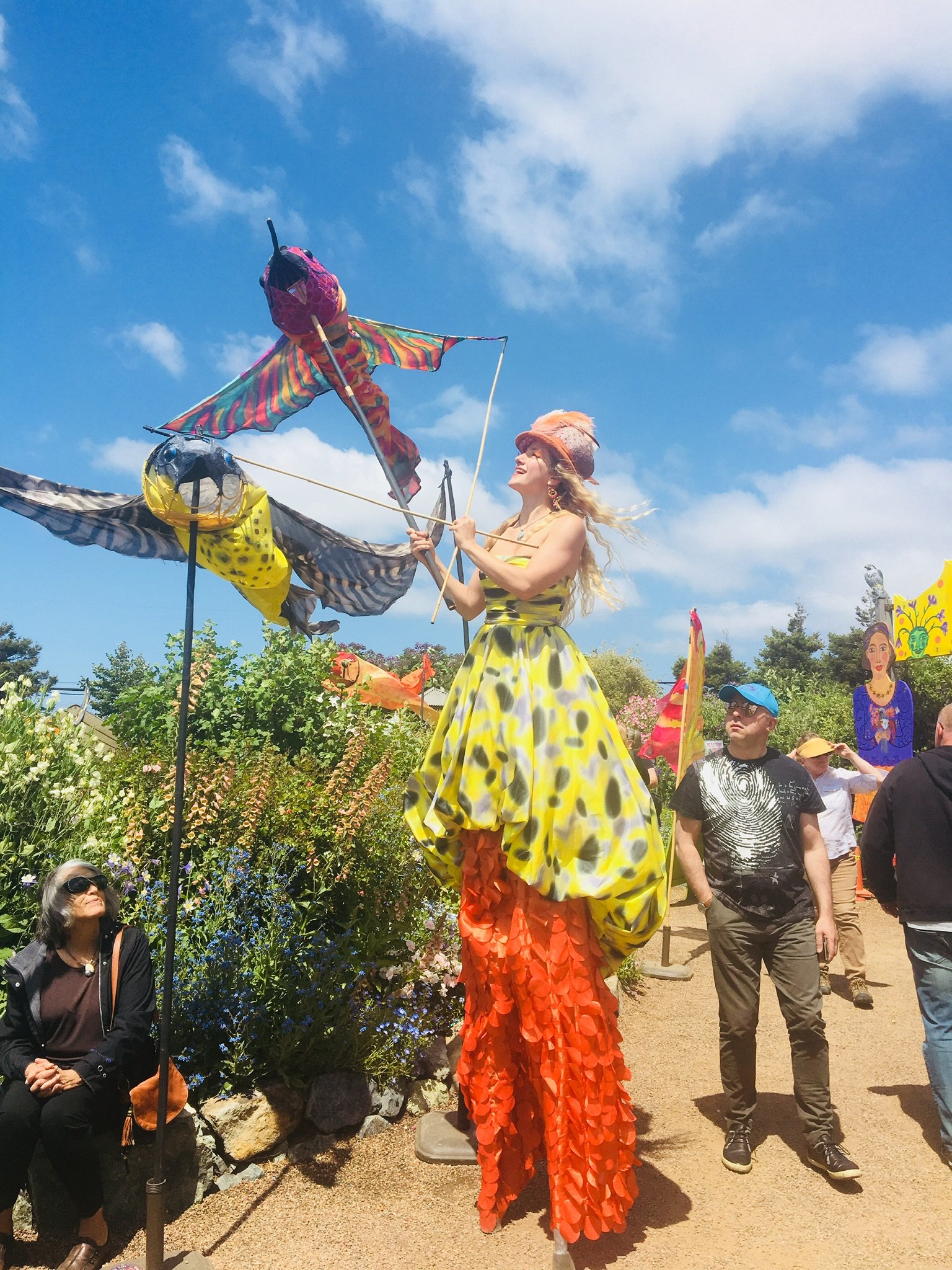 Colorful outdoor scene featuring a large human-like puppet with a yellow and black dress and orange textured legs, operating a kite-like object, surrounded by people and greenery on a sunny day.