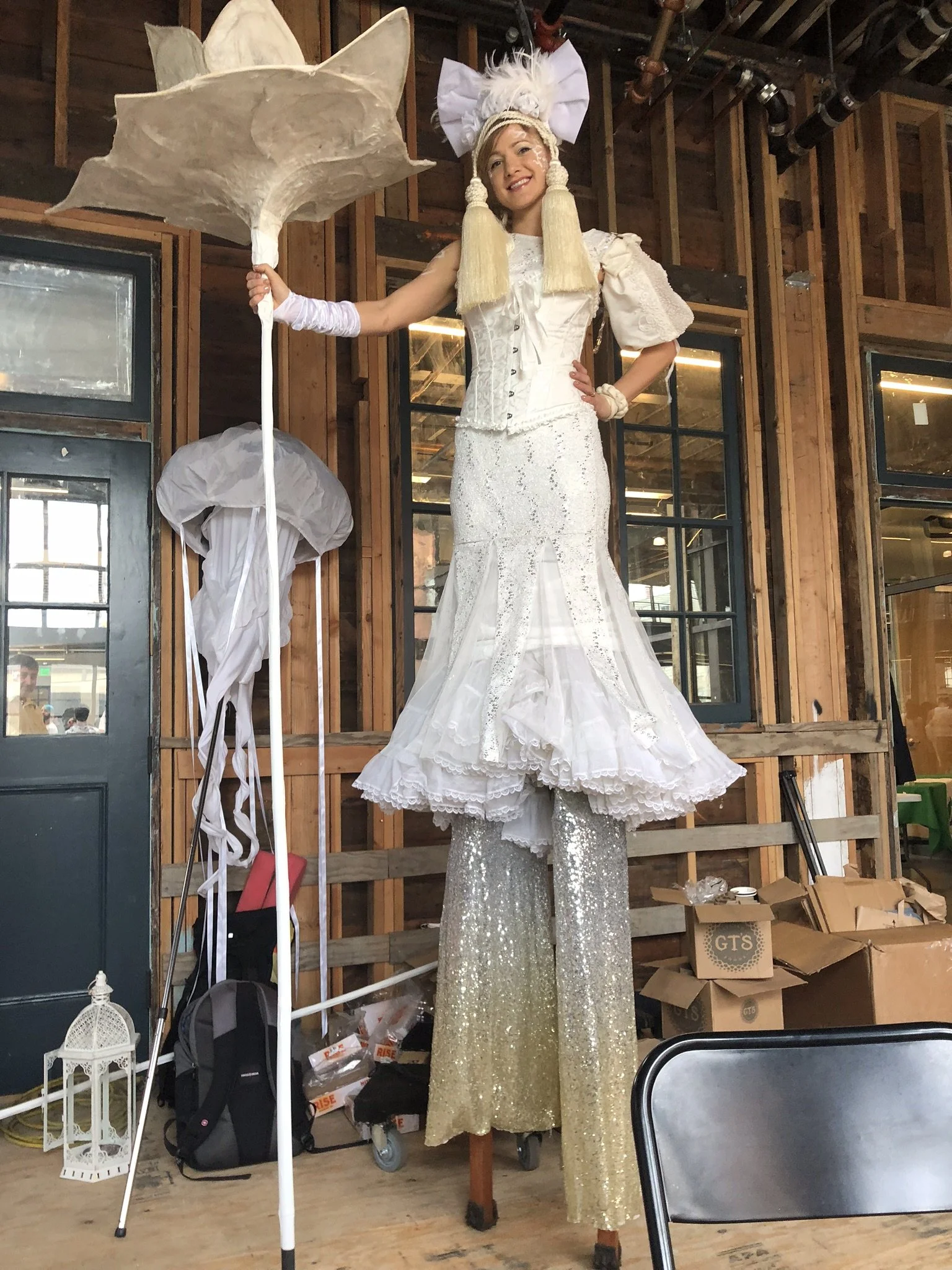 A woman on stilts dressed in an elaborate, all-white costume with a lace top, ruffled skirt, and sparkly pants. She wears a large headpiece with white pom-poms and long blonde tassels, and she is smiling while holding a large, decorative white paraso