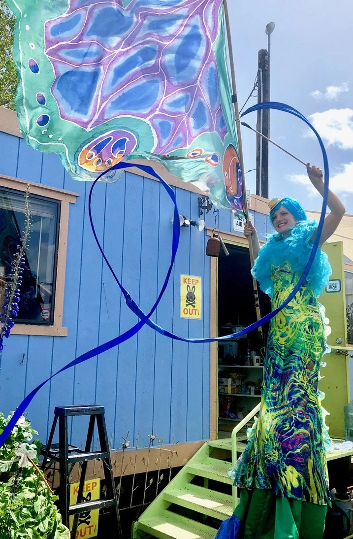 A person with blue hair in a colorful tie-dye dress is standing on steps outside a blue building, flying a large, colorful butterfly-shaped kite with purple, blue, green, and orange patterns.