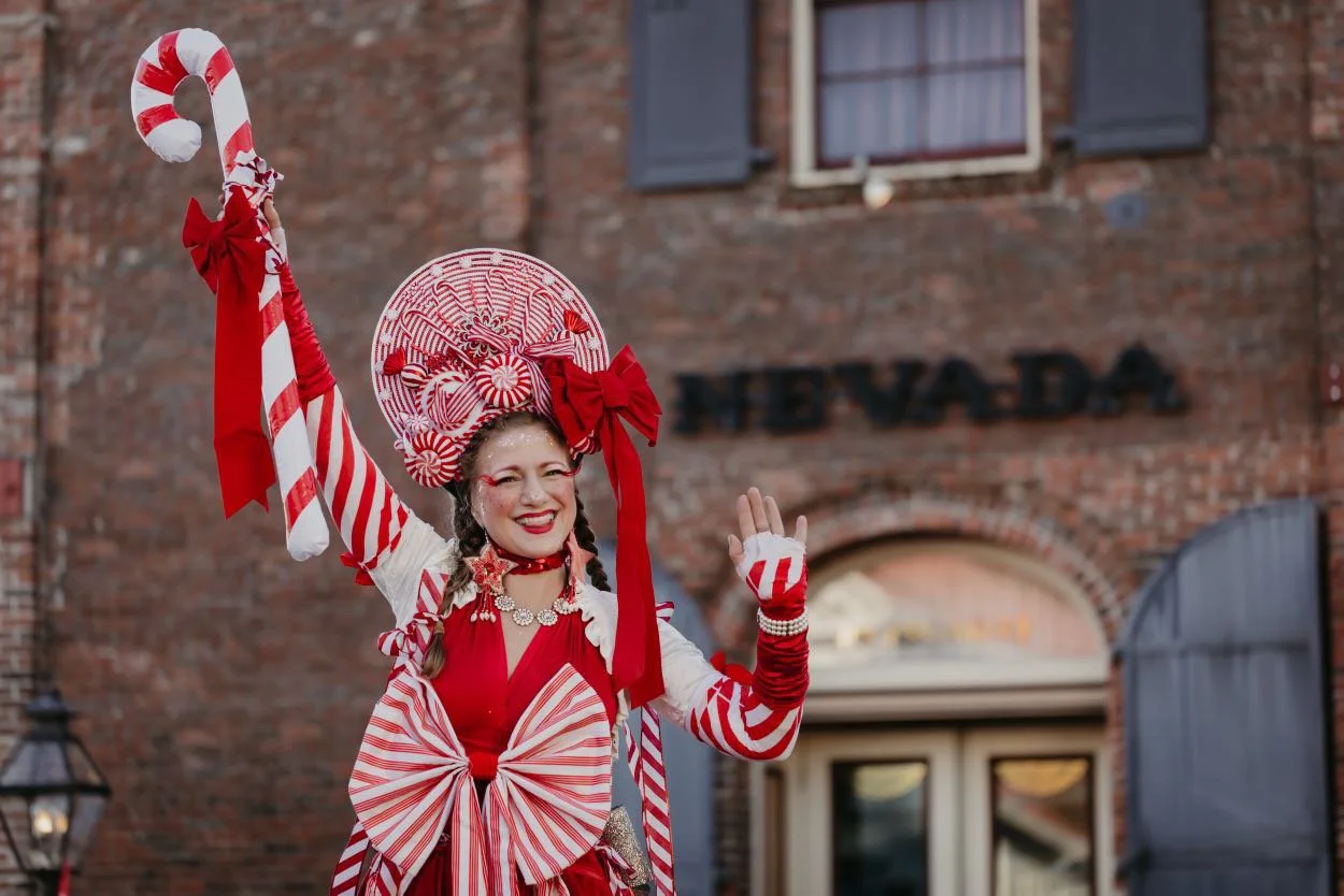 Woman dressed in a red and white candy-themed costume, holding a candy cane prop, smiling, in front of a brick building with the sign 'N E V A D A' behind her.